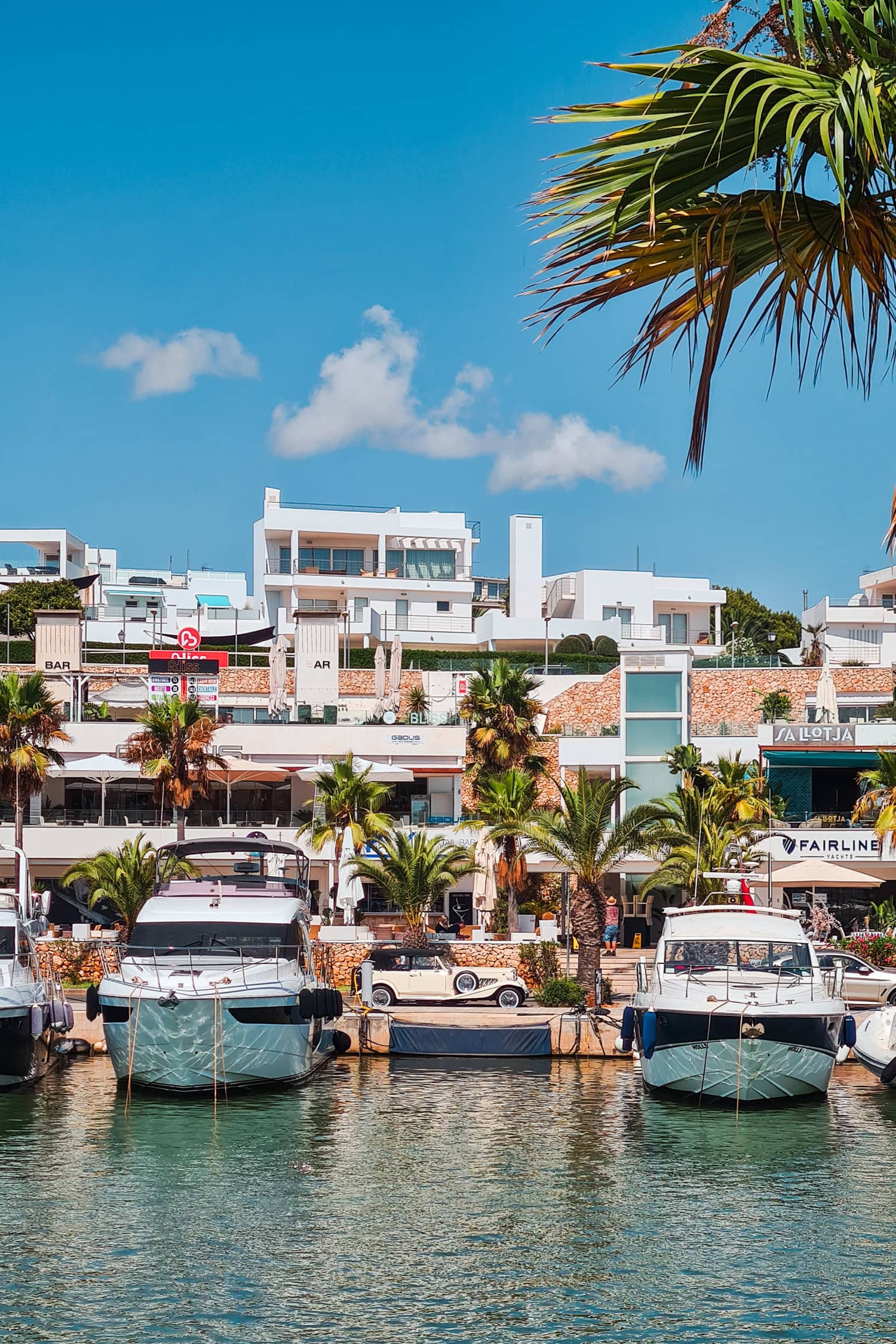 Two large black and white boats with a vintage beige car in the background surrounded by palms in Cala d'Or Mallorca.