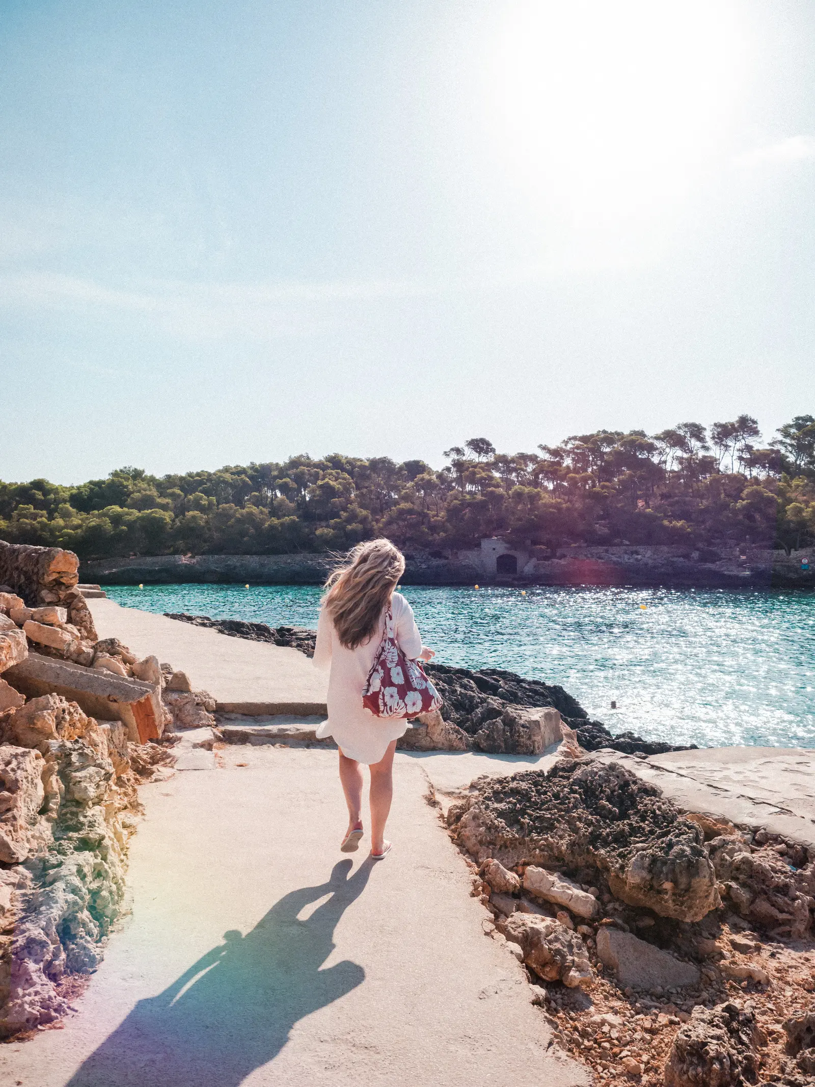 Woman in a white dress, carrying a burgundy bag, walking along a path next to turquoise water in Cala Mondrago Mallorca.