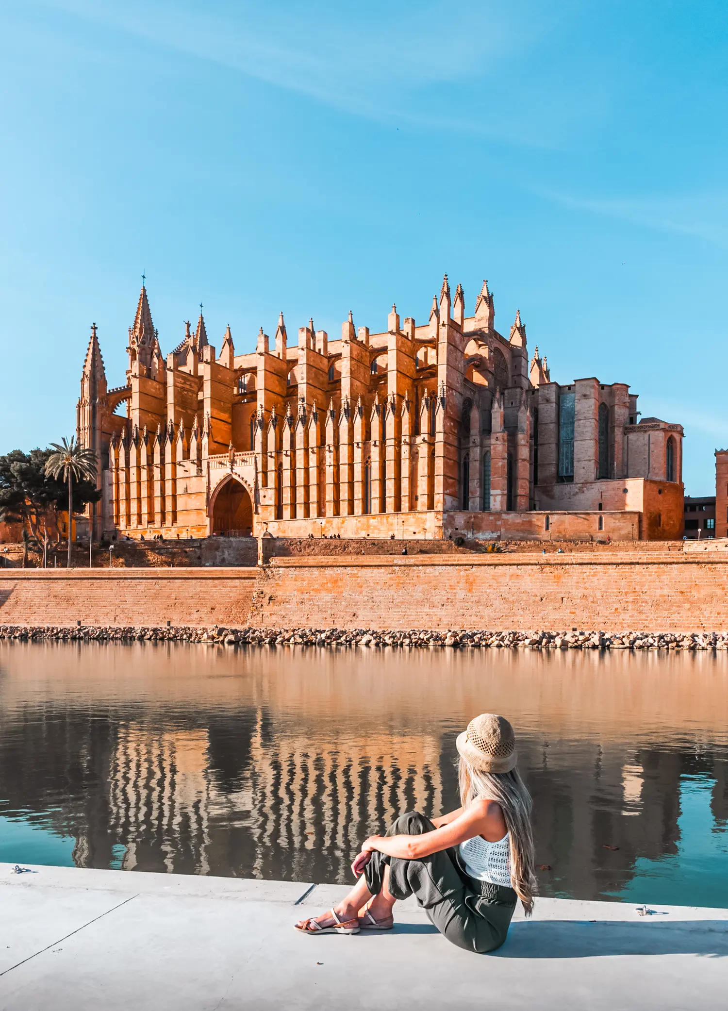 Girl with long hair, wearing green pants and a white top, sitting along the river in Palma de Mallorca looking over at the beige Palma Cathedral on the other side.