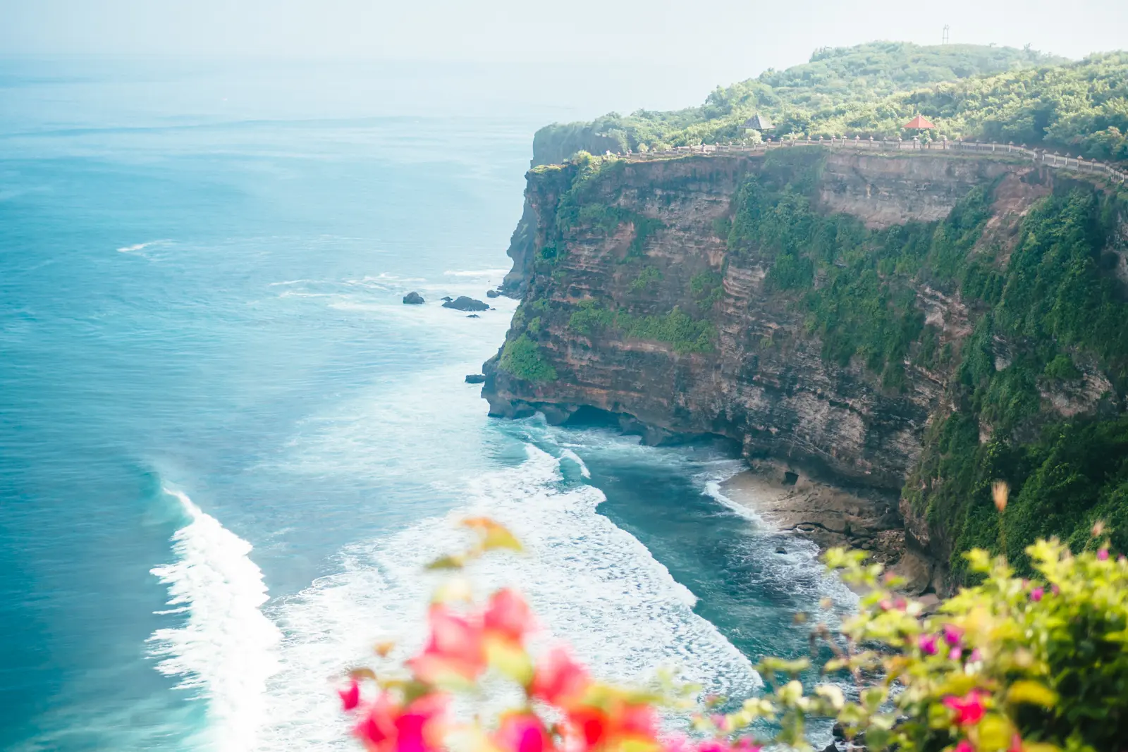 View from Uluwatu Temple in Bali over the ocean and surrounding cliffs with red flowers in the foreground.