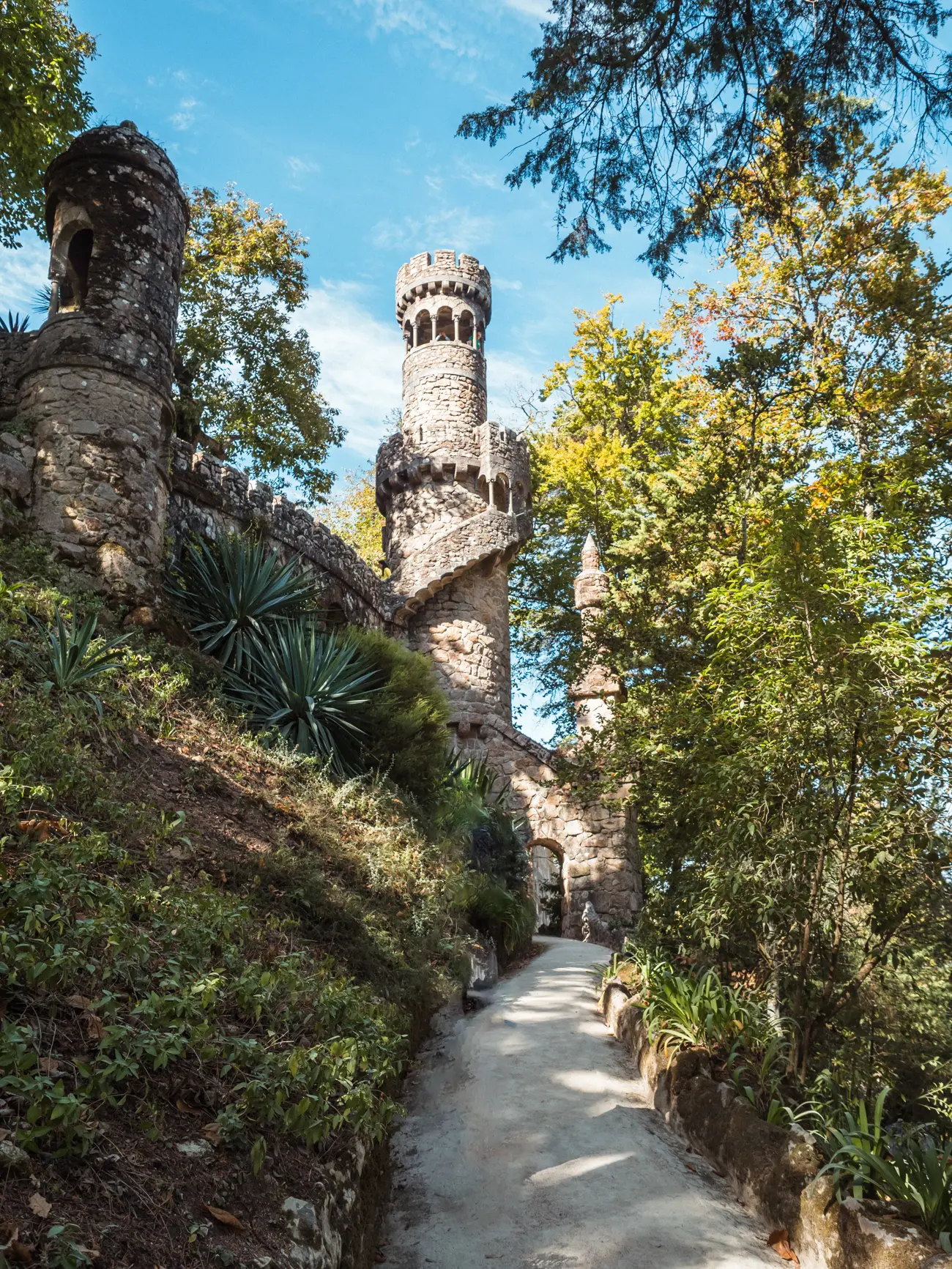 Path leading towards narrow light grey stone tower in the forest at Quinta da Regaleira, the best palace in Sintra Portugal.