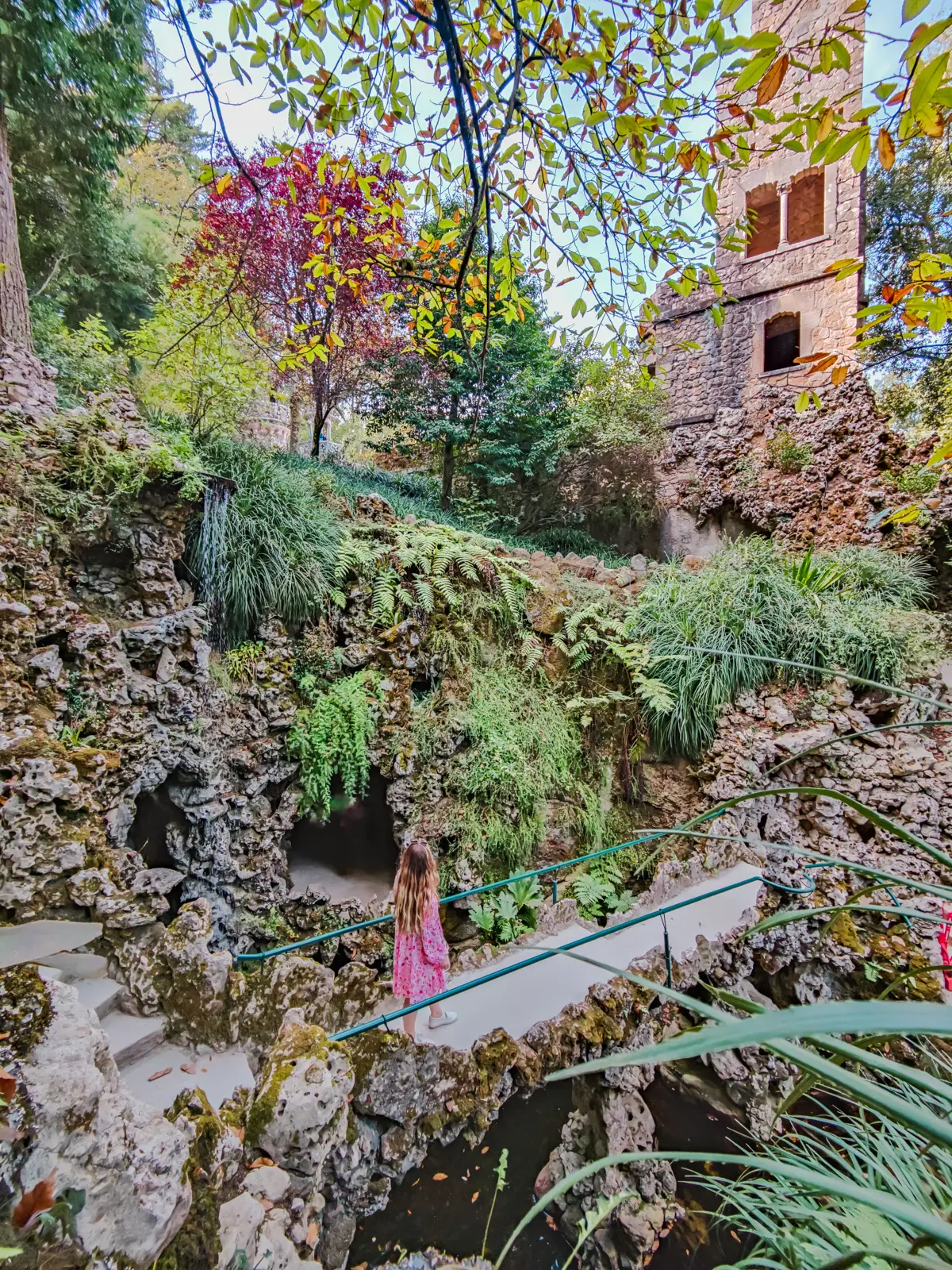 Girl in a pink dress and white shoes walking over a stone bridge along a stone wall with green plants at Quinta da Regaleira palace in Sintra Portugal.