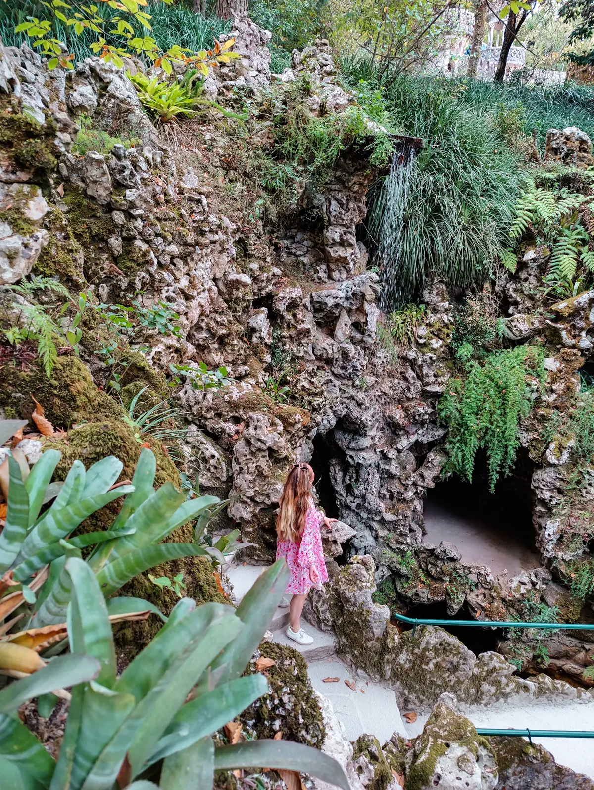 Girl in a pink dress and white shoes walking down grey stone stairs along a stone wall with green plants at Quinta da Regaleira palace in Sintra Portugal.