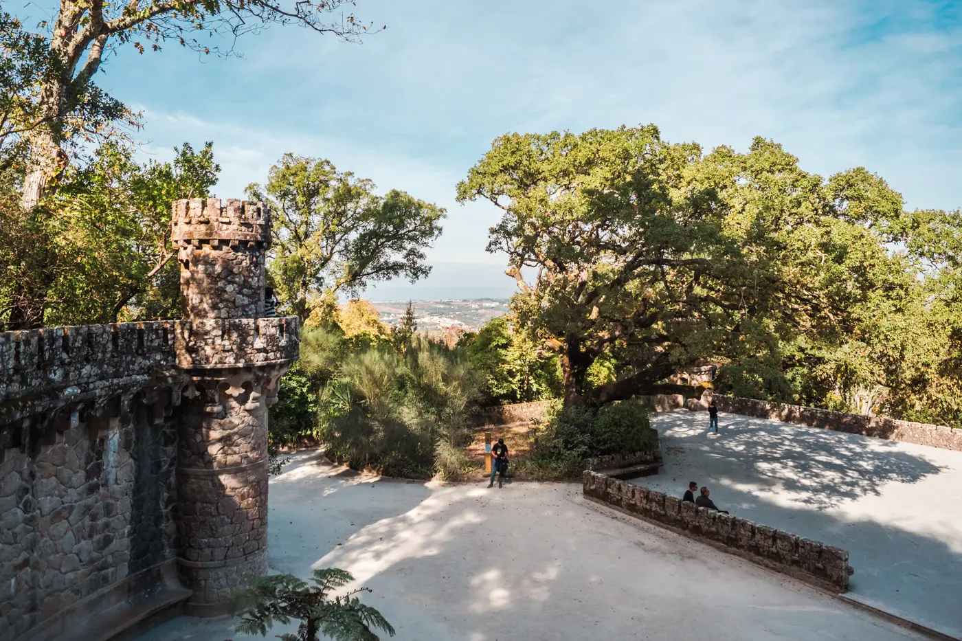 Medival looking stone wall with small tower to the left, open area with light grey gravel surrounded by green trees at Quinta da Regaleira, the most beautiful palace in Sintra.