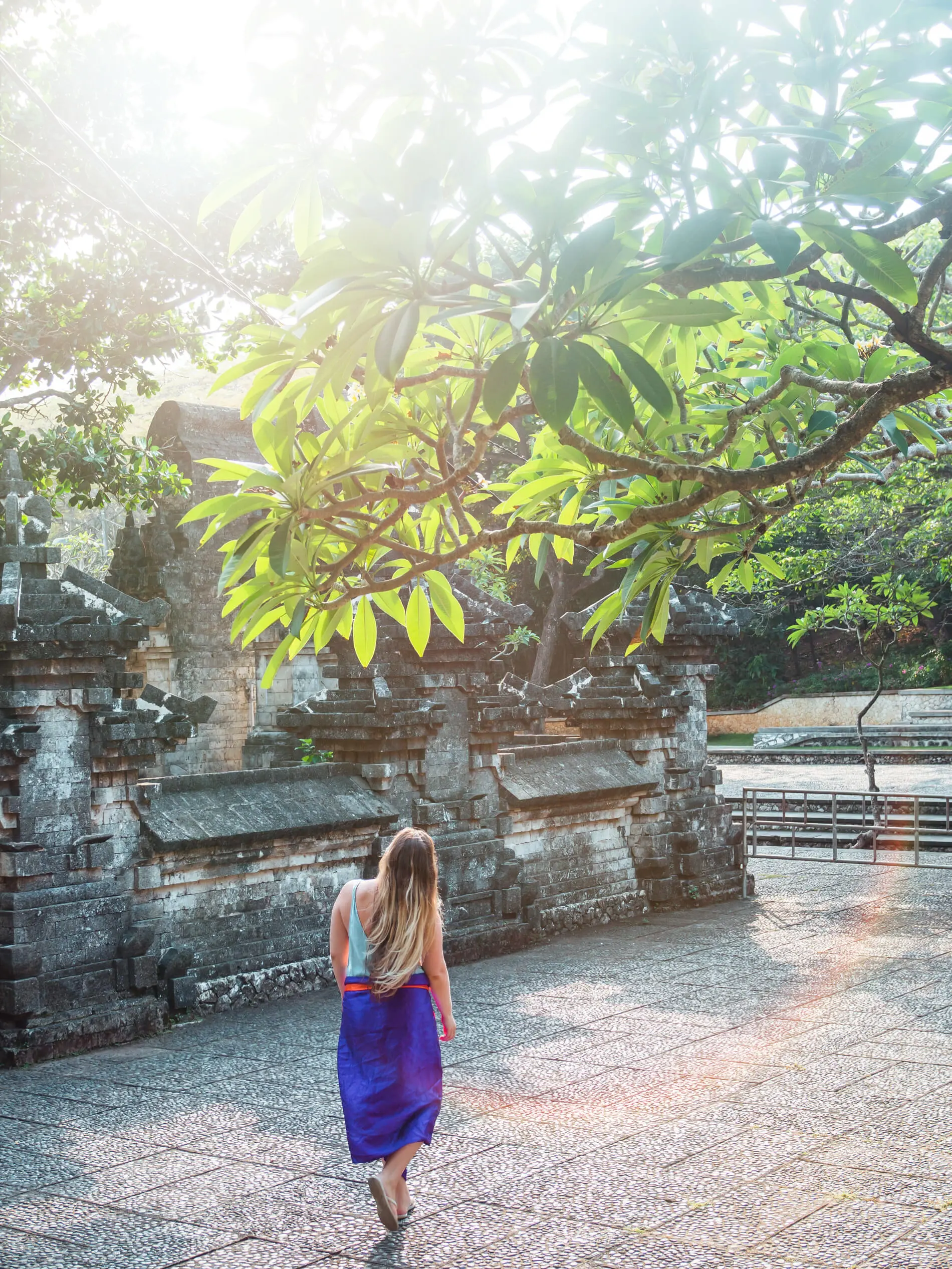 Girl with long hair walking along an ancient ornate shrine in white and grey stone under a tree with sun shining in at Uluwatu Temple in Bali.