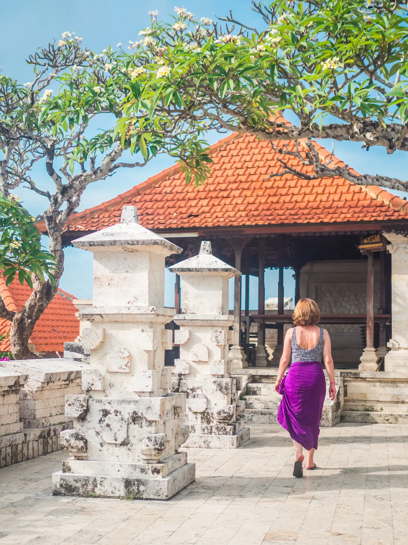 Woman wearing a purple sarong walking past white pillars towards an altar with orange roof at Uluwatu Temple.
