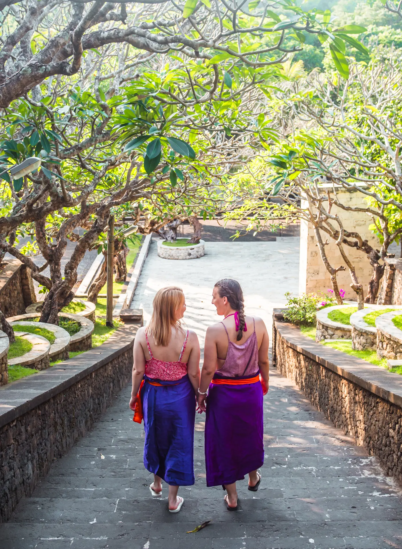 Two girls wearing purple sarongs walking down the stone stairs at Uluwatu Temple under a canopy of green leaves.