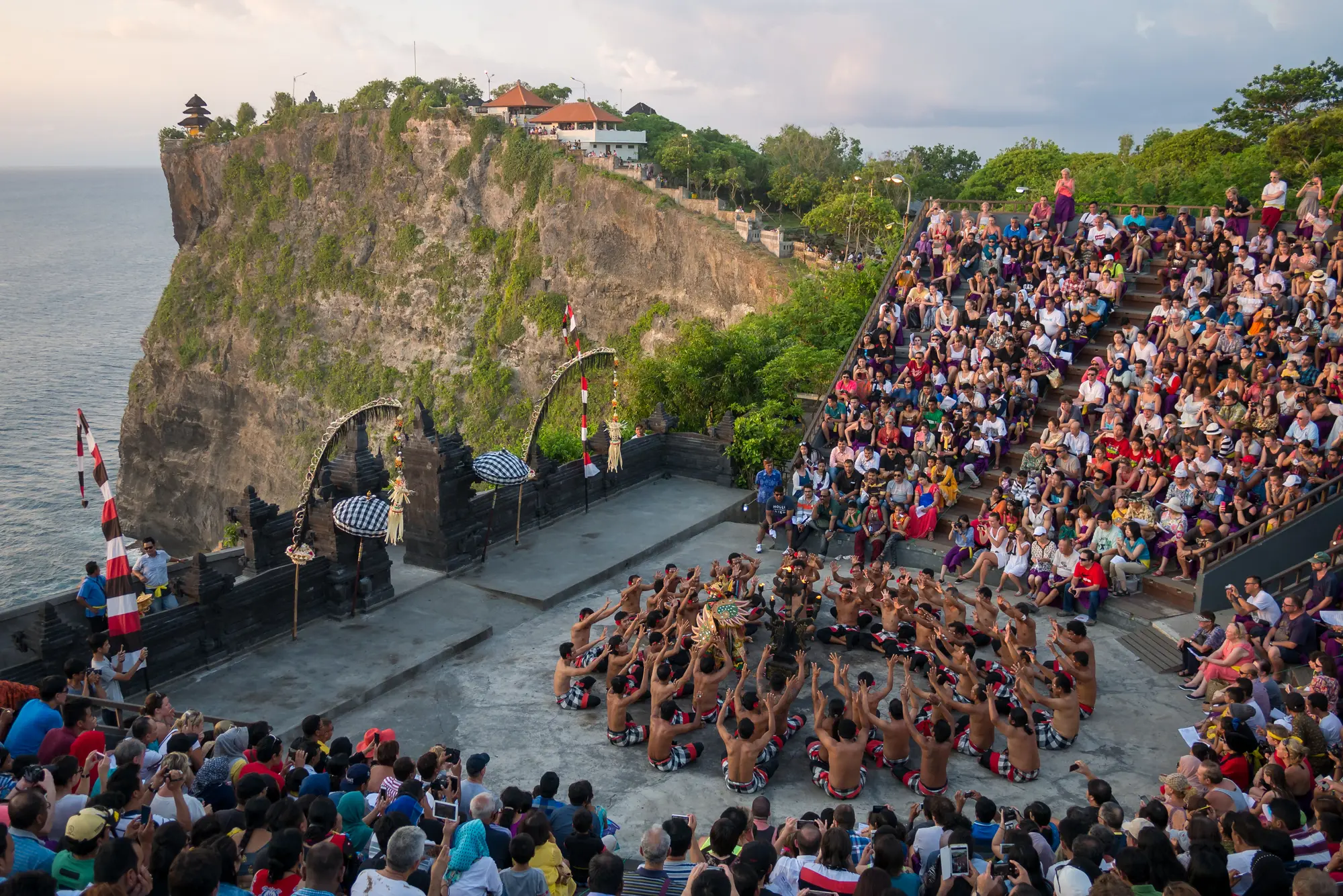 Amphitheater full of people set on the edge of Uluwatu Temple with a cliff in the background at sunset looking at a ring of Kecak dancers.