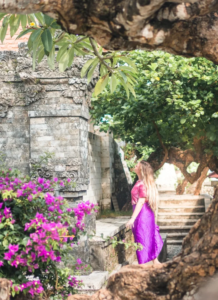 Girl with long hair wearing a purple sarong looking at a grey stone temple in Uluwatu surrounded by purple flowers and green trees.