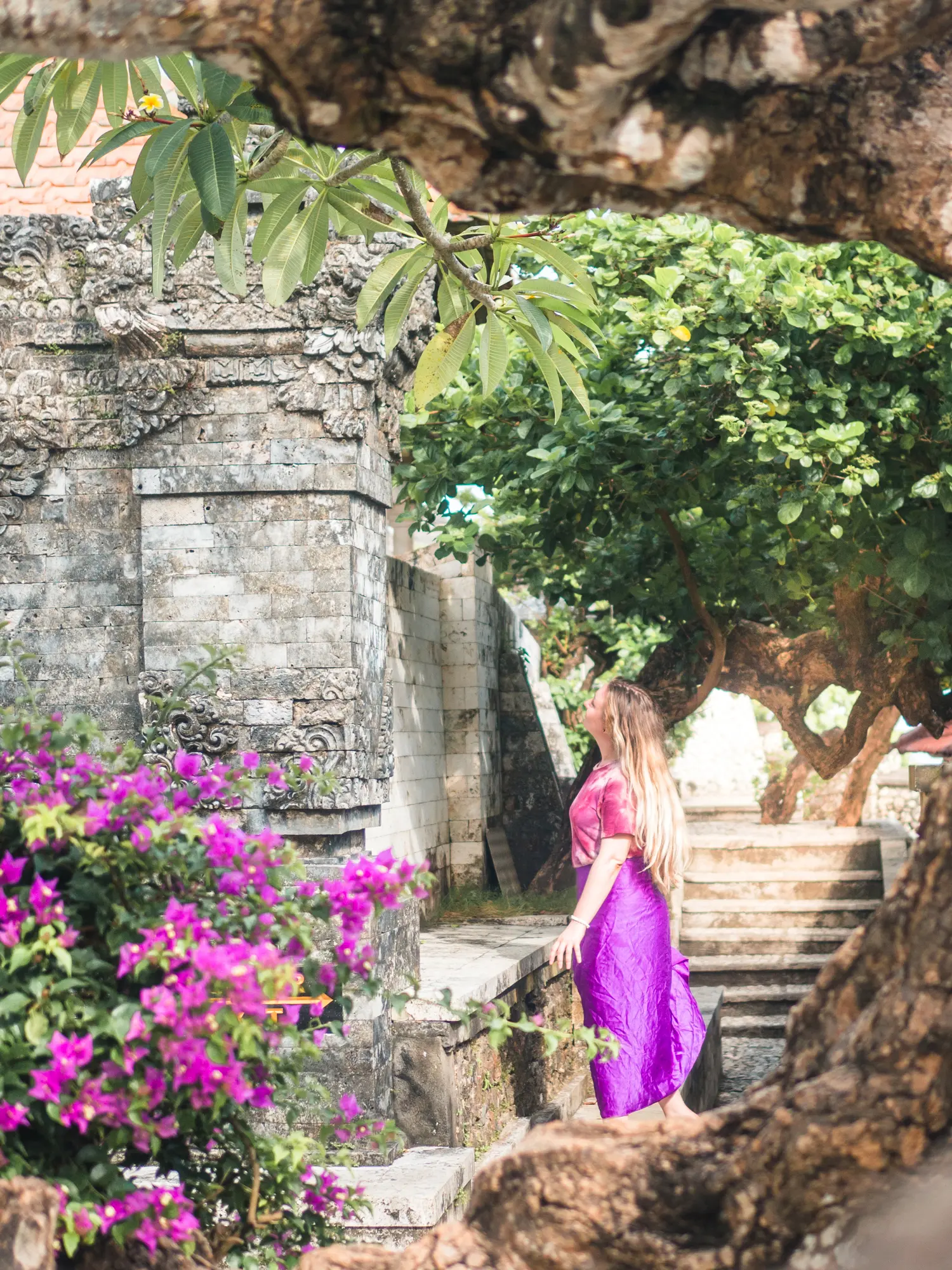 Girl with long hair wearing a purple sarong looking at a grey stone temple in Uluwatu surrounded by purple flowers and green trees.