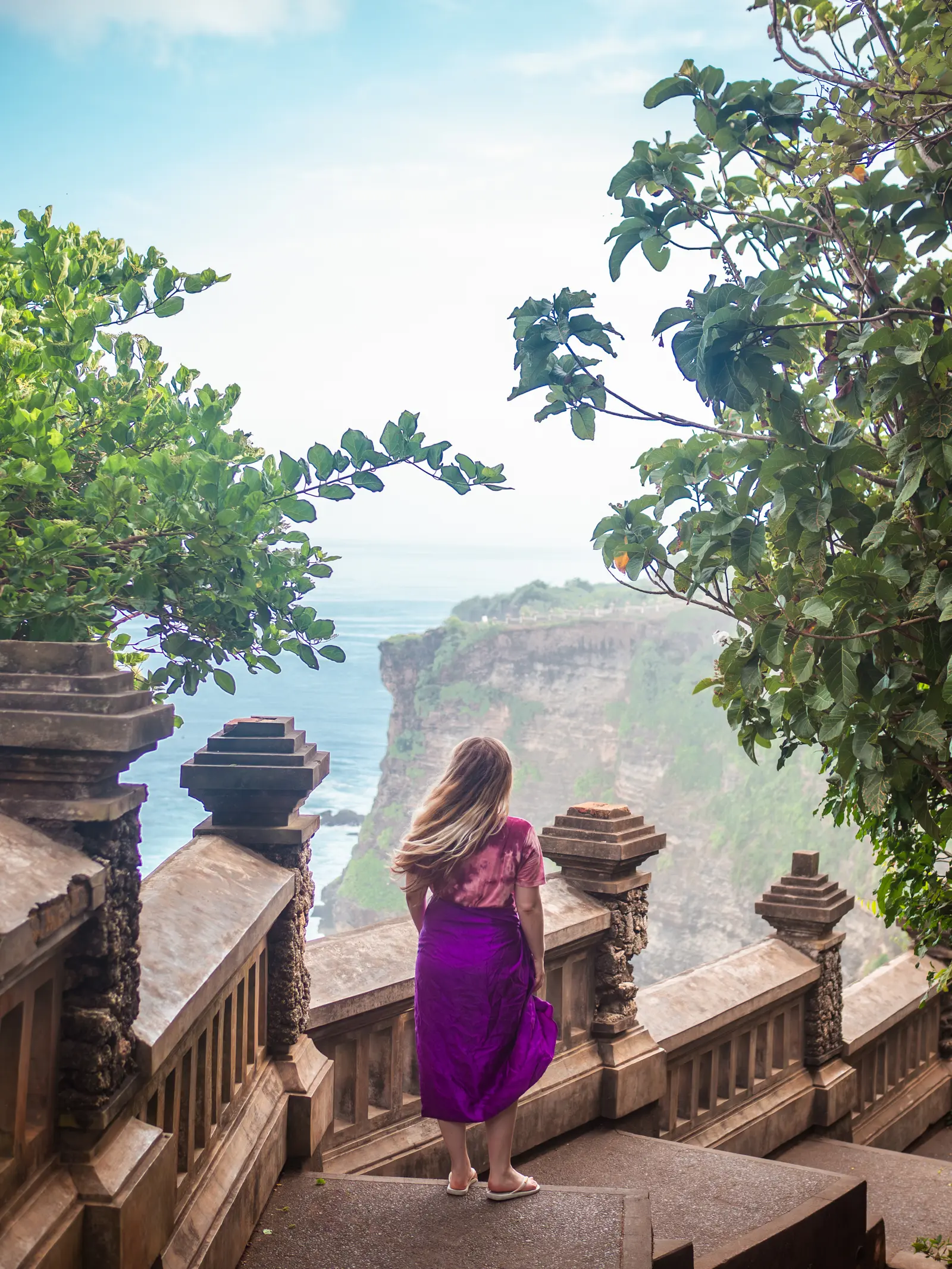 Girl with long hair wearing purple sarong walking down stairs on the edge of Uluwatu Temple in Bali looking out over the ocean and cliffs.