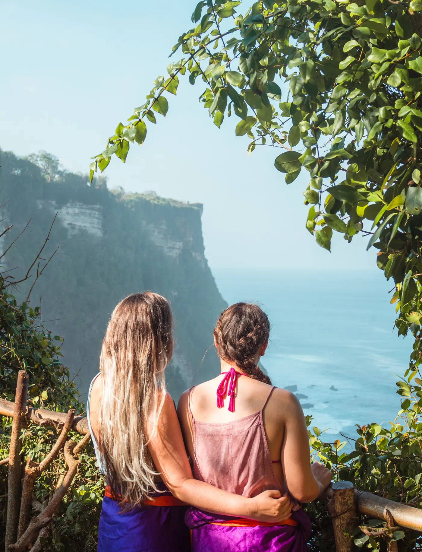 Girl with long hear wearing a blue sarong standing next to a girl wearing a pink sarong looking out over the cliffs form Uluwatu Temple.