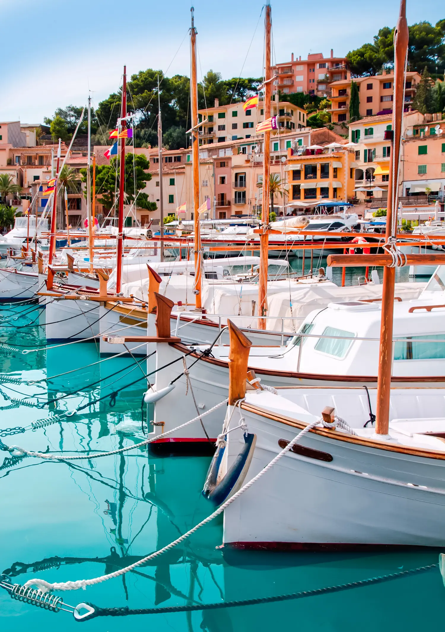 Mallorca - White sailboats in a row in the turquoise water in Port de Sóller with beige buildings in the background.