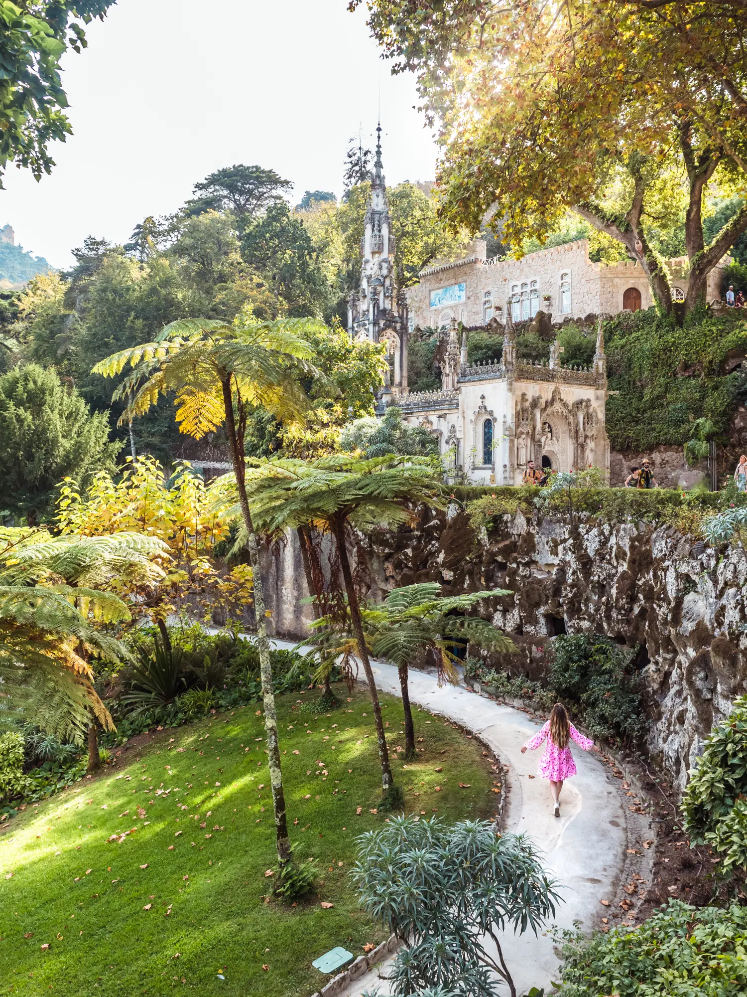 Girl with long hair, n a pink dress, walking on a light grey path along the stone wall of Quinta da Regaleira palace surrounded by green grass and trees.