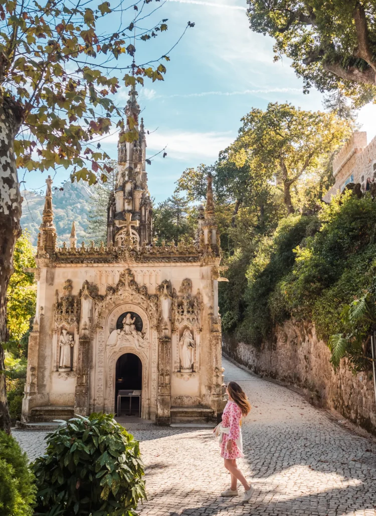 Girl with long dark blonde hair, wearing a pink dress and white shoes, walking towards the ornate chapel on the grounds of Quinta da Regaleira in Sintra, surrounded by green gardens and a cobbled walkway.