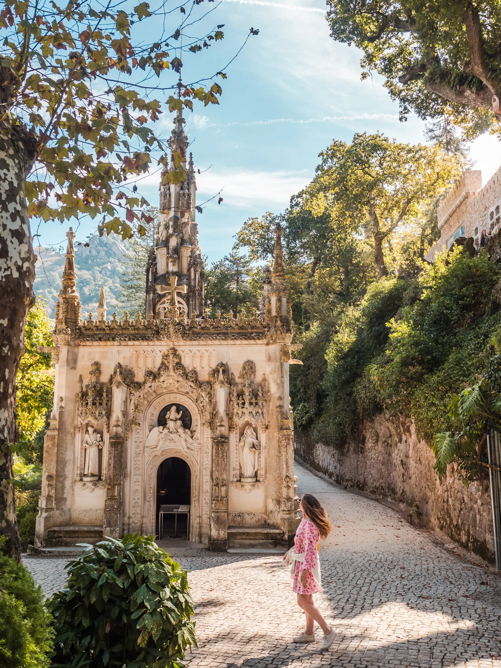 Girl with long dark blonde hair, wearing a pink dress and white shoes, walking towards the ornate chapel on the grounds of Quinta da Regaleira in Sintra, surrounded by green gardens and a cobbled walkway.