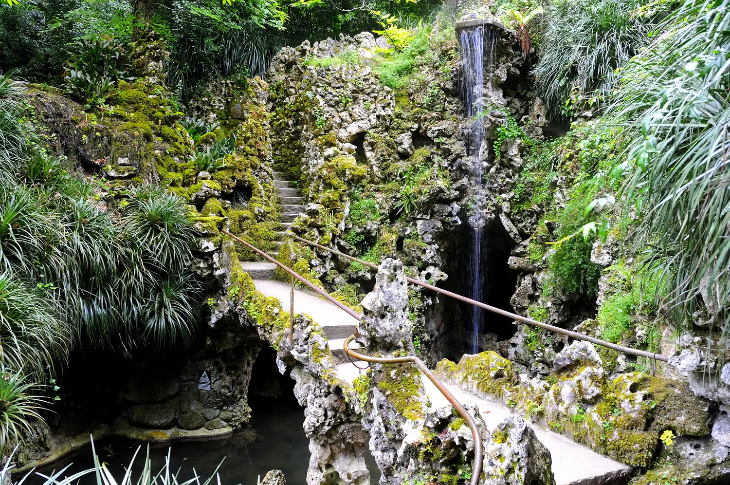 Grey narrow stone bridge with moss leading towards a staircase surrounded by a greenery and a small waterfall on the left at Quinta da Regaleira in Sintra.