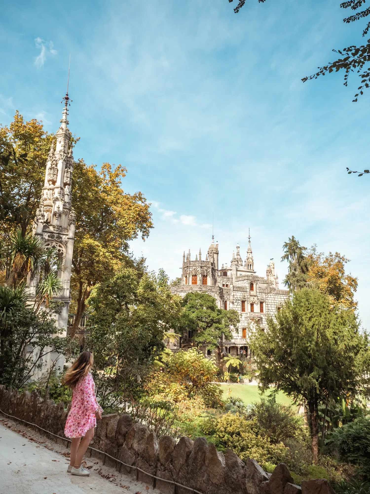 Girl with long dark blonde hair, in a pink dress, standing in front of the lush gardens of Quinta da Regaleira in Sintra, looking towards a 19th century grey stone palace with spires.