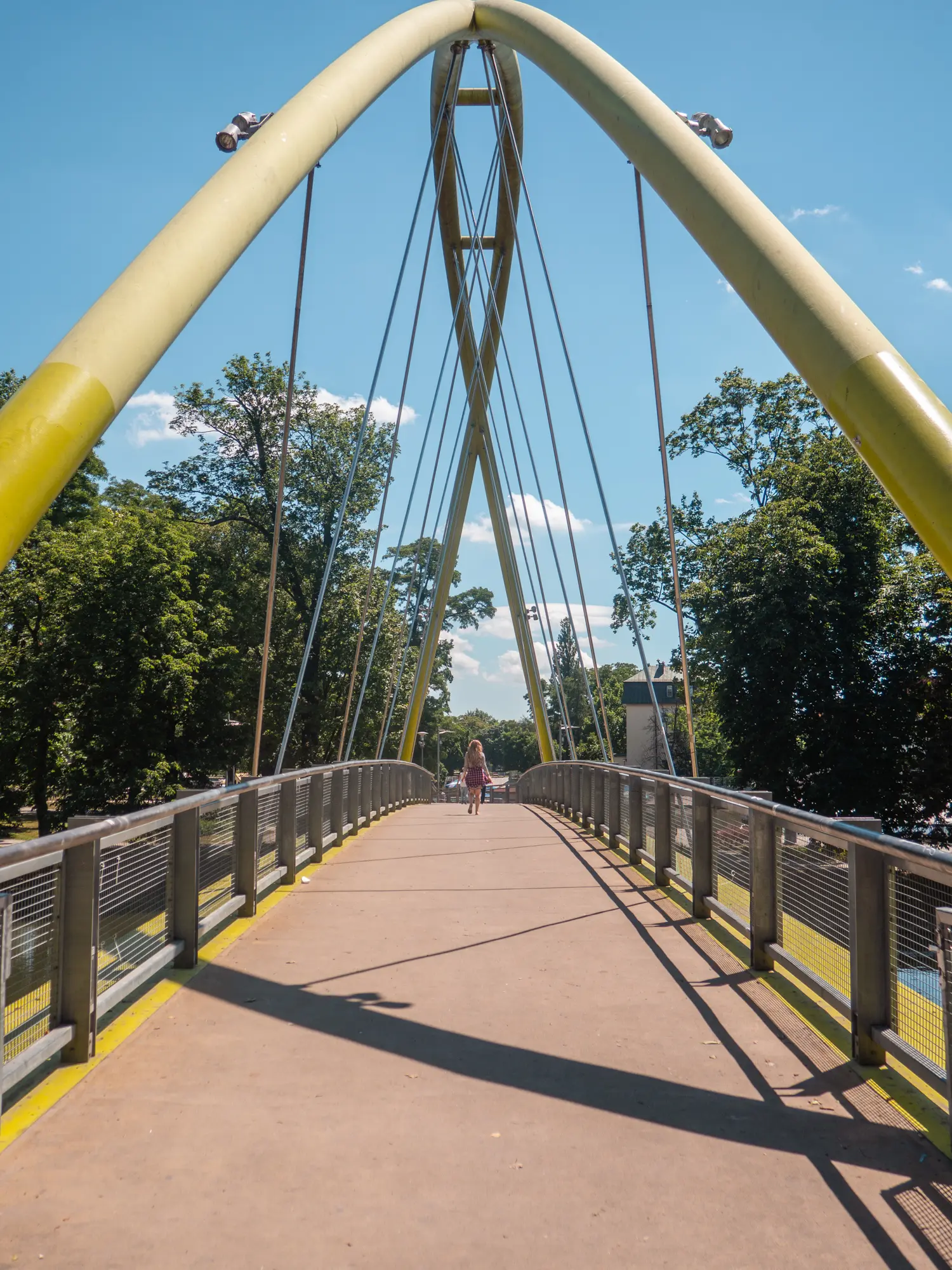 Girl walking over yellow modern bridge surrounded by green trees on a sunny day in Wroclaw.