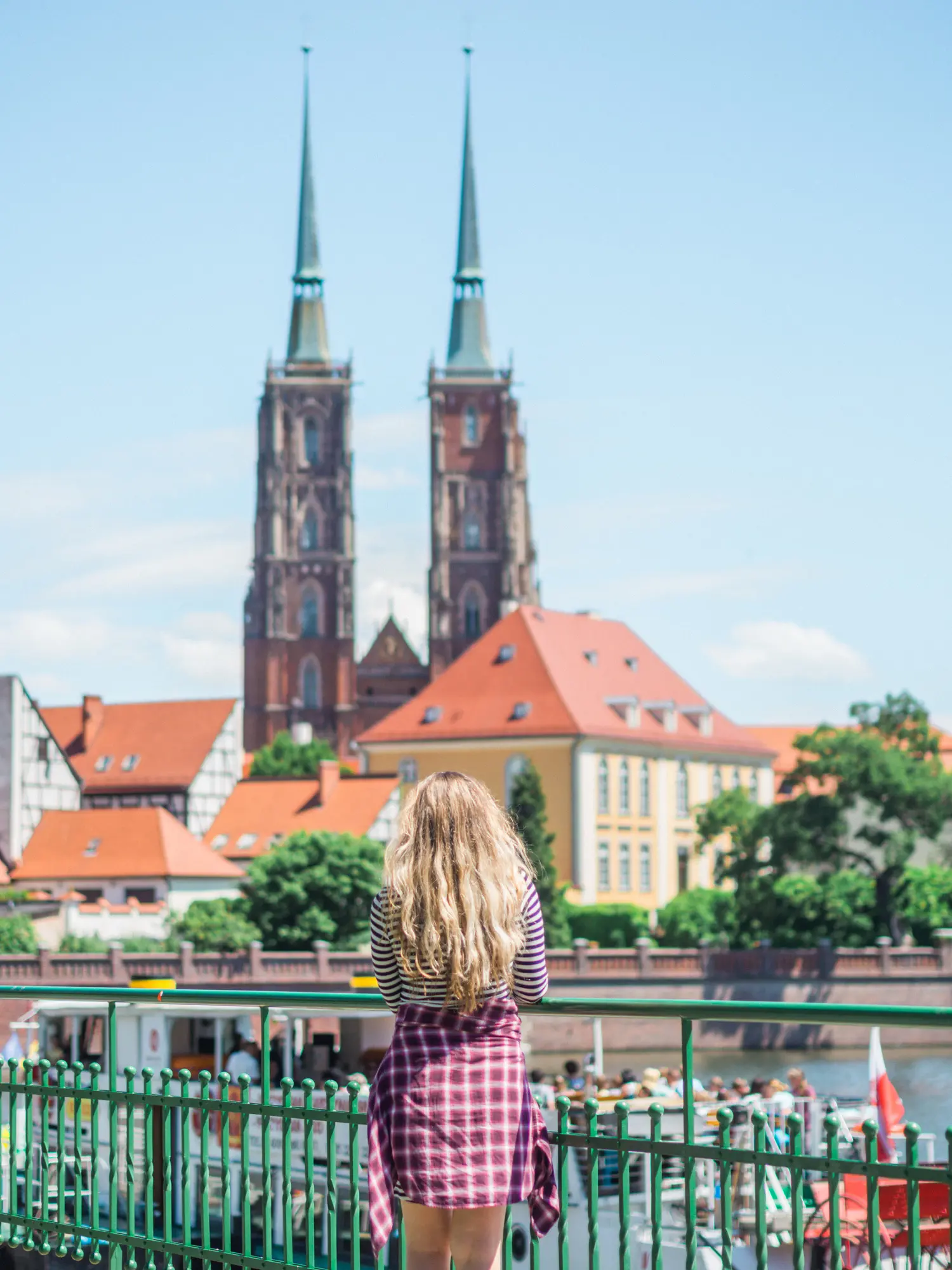 Girl with long blonde hair, wearing a purple shirt, standing on a green metal bridge looking over at Wroclaw Cathedral.