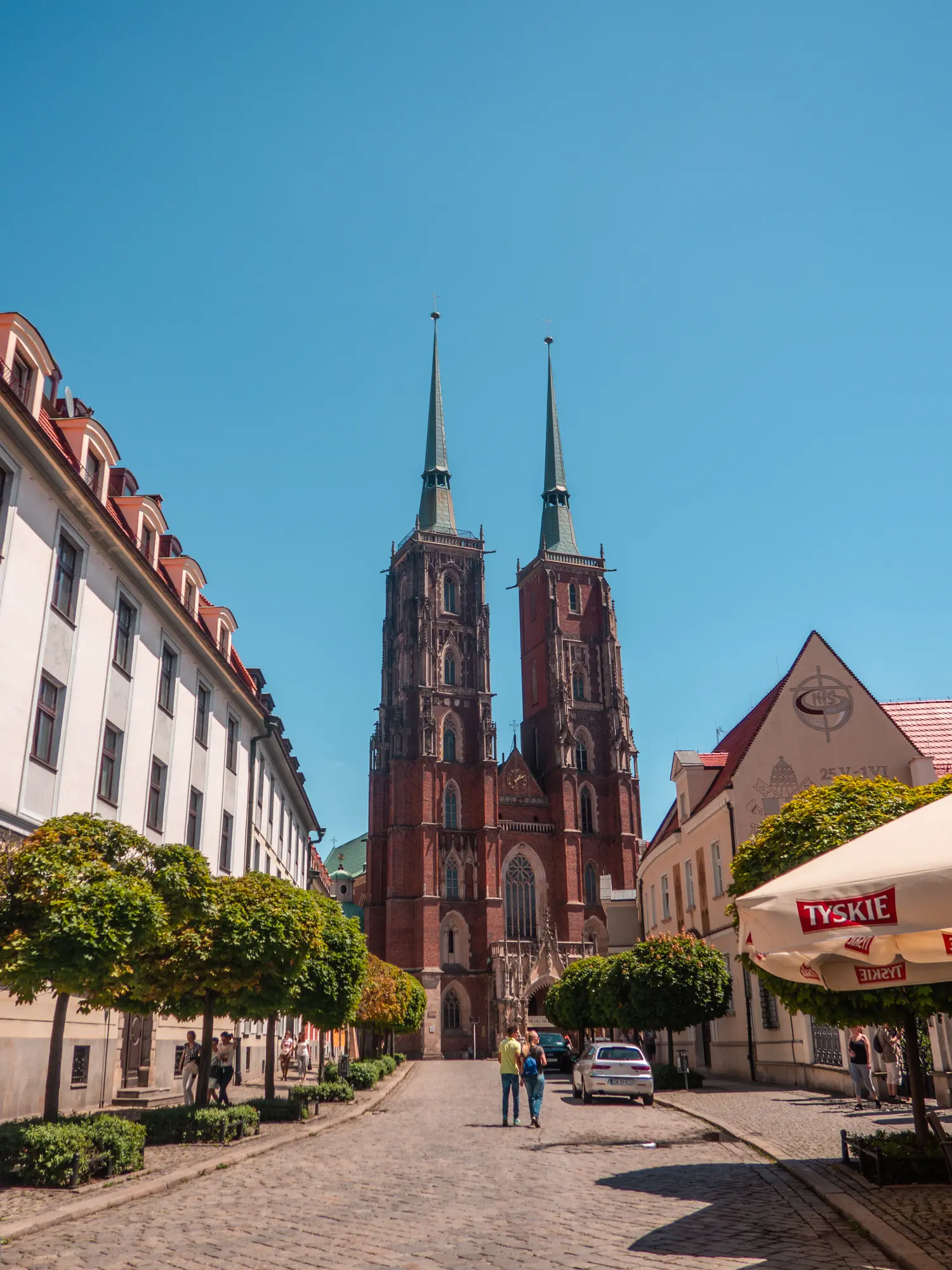 Wroclaw Cathedral, a large brick church with two spiers, old buildings on both sides, on a sunny day.
