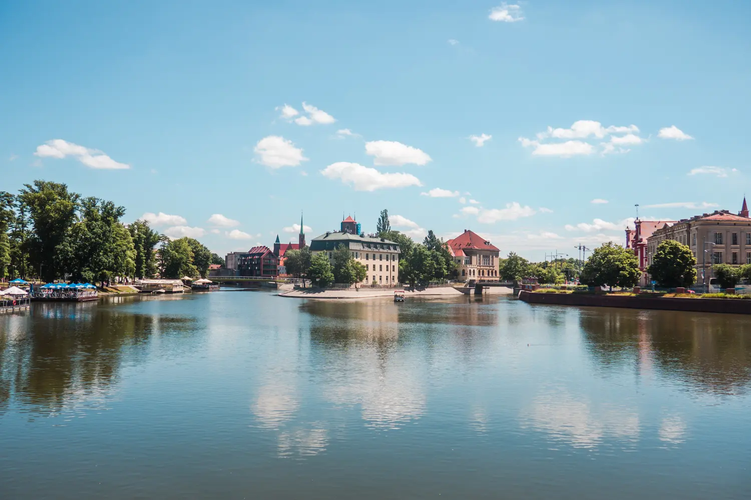 View of the clam, blue river in Wroclaw, with green trees and old building in the background.