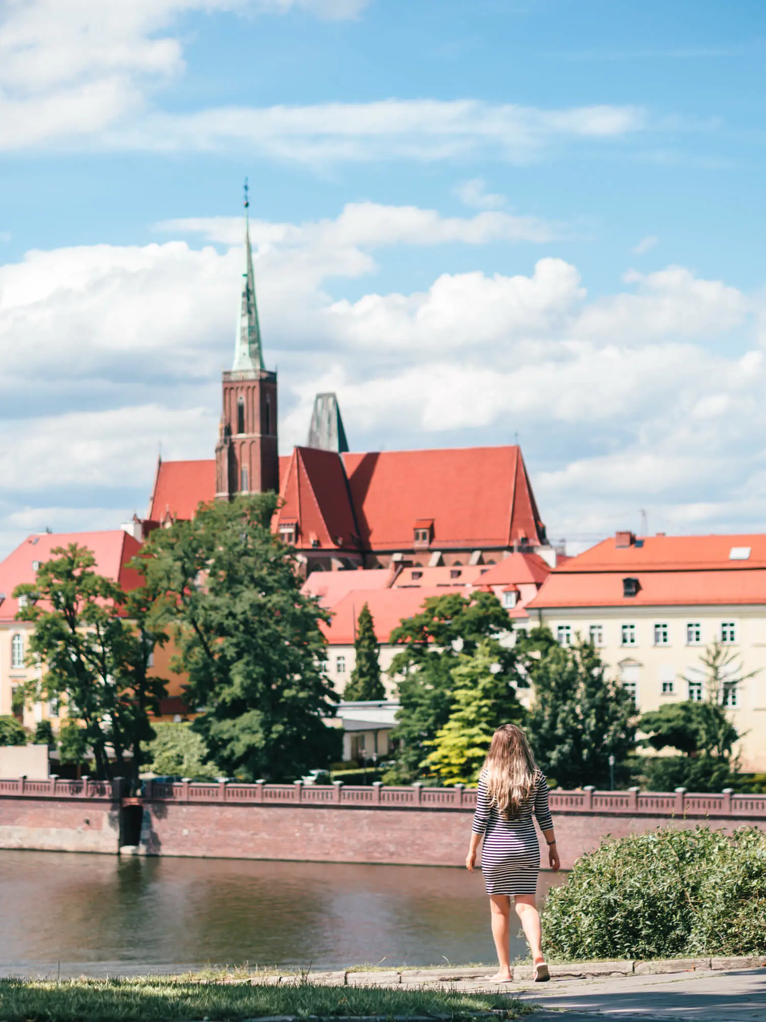Girl wearing a striped dress, walking down towards the river in Wroclaw with old buildings and a church on the other side.