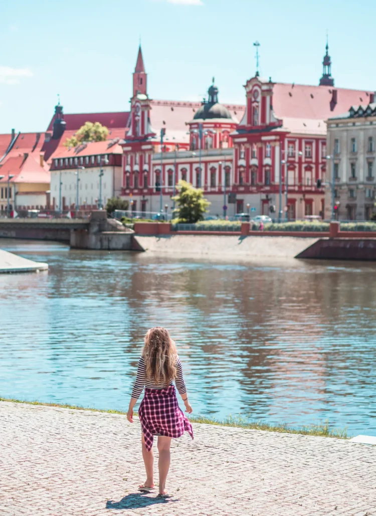 Girl with long hair, wearing a striped dress and purple shirt, walking along the river in Wroclaw with red old buildings in the background.