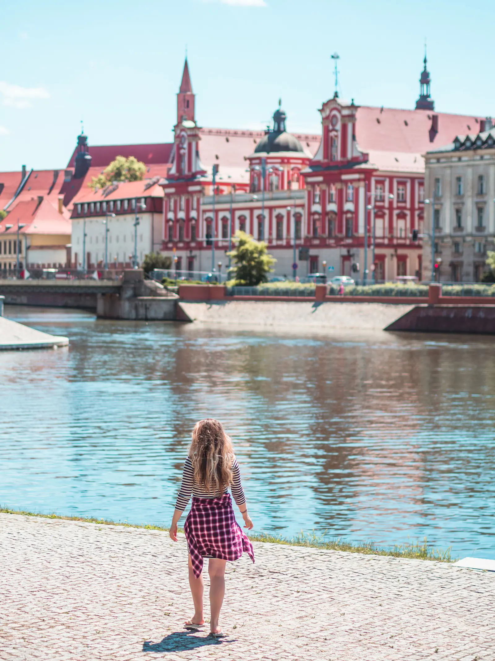 Girl with long hear, wearing a striped dress and purple shirt, walking along the river in Wroclaw with red old buildings in the background.