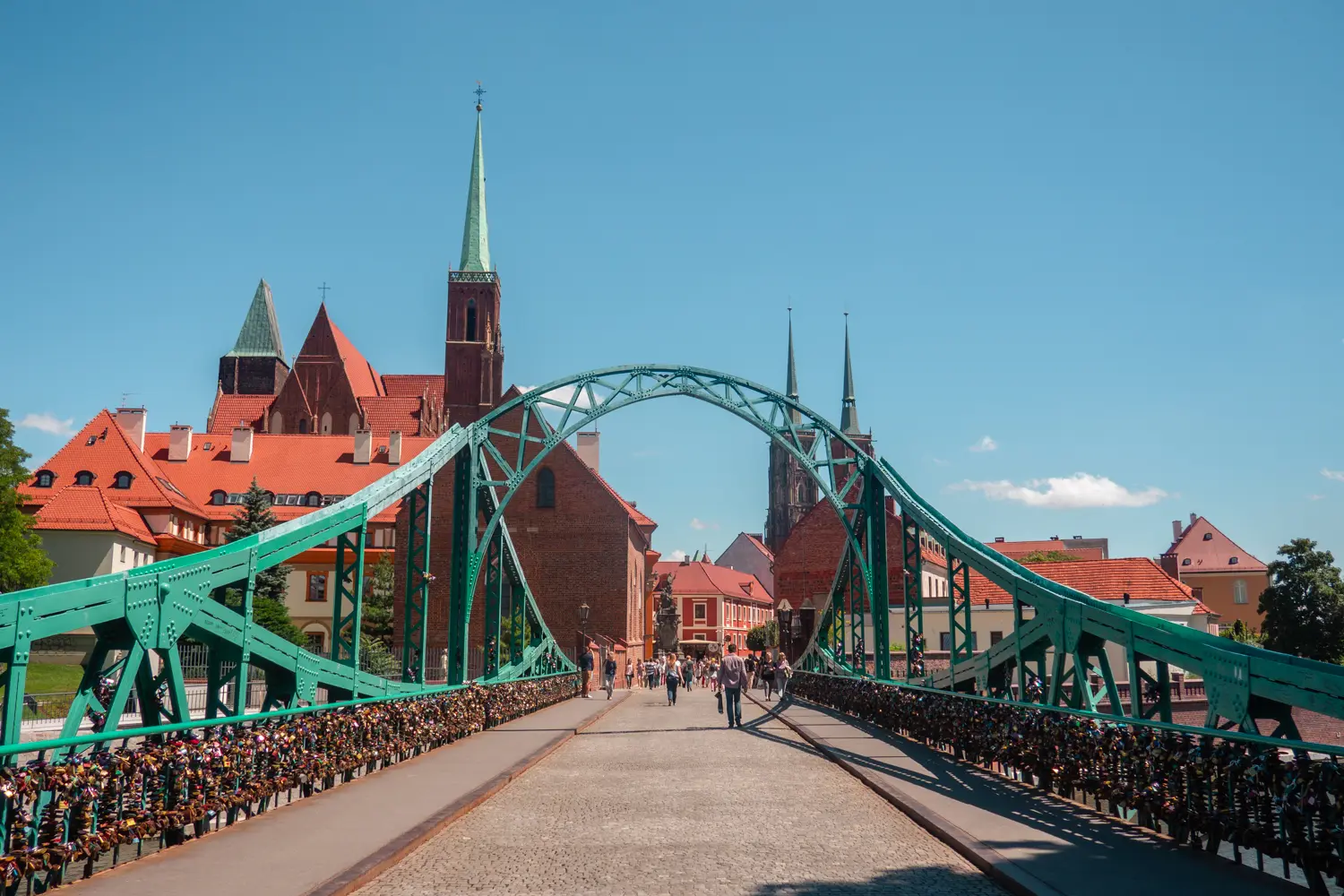 Green metal bridge with love locks on both sides, two brick churches in the background, in Old Town Wroclaw.