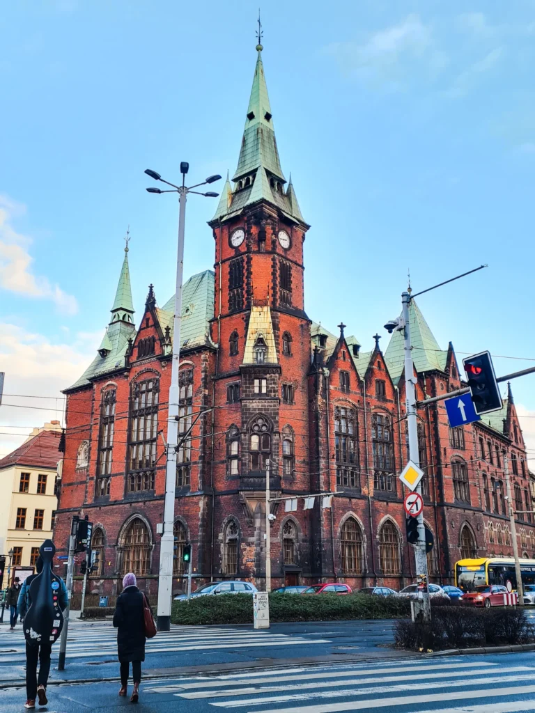 Looking across the street at a Gothic brick building with copper ceiling in the towers, housing Wroclaw University Library.