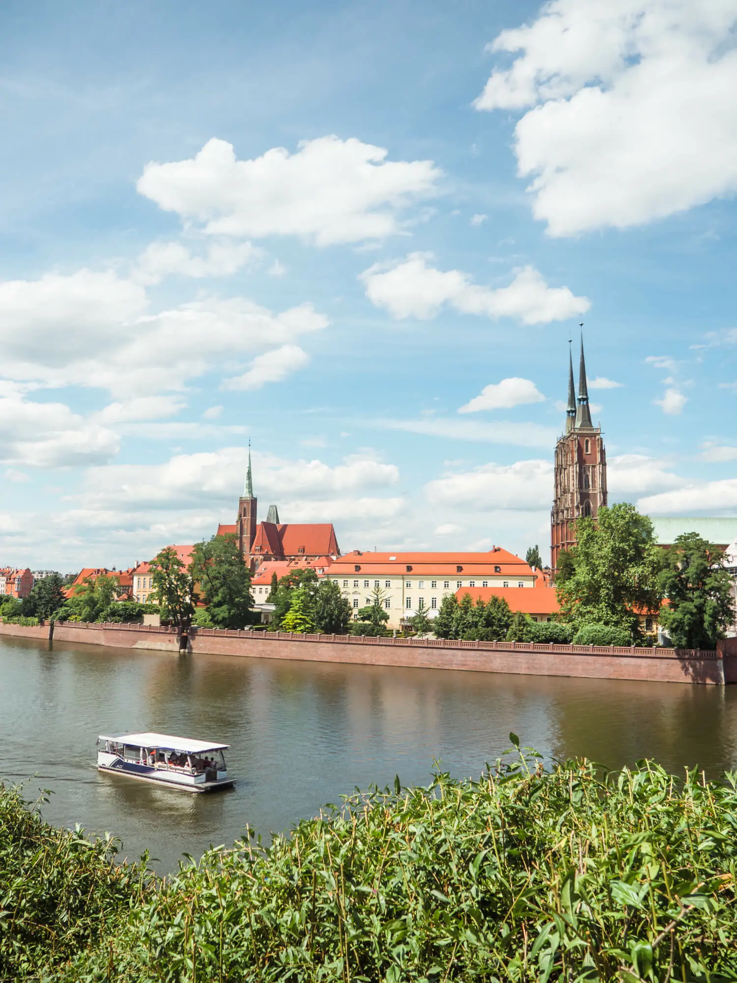 View of boat on Wroclaw river lined with greenery, with two church spires and old buildings in the background.