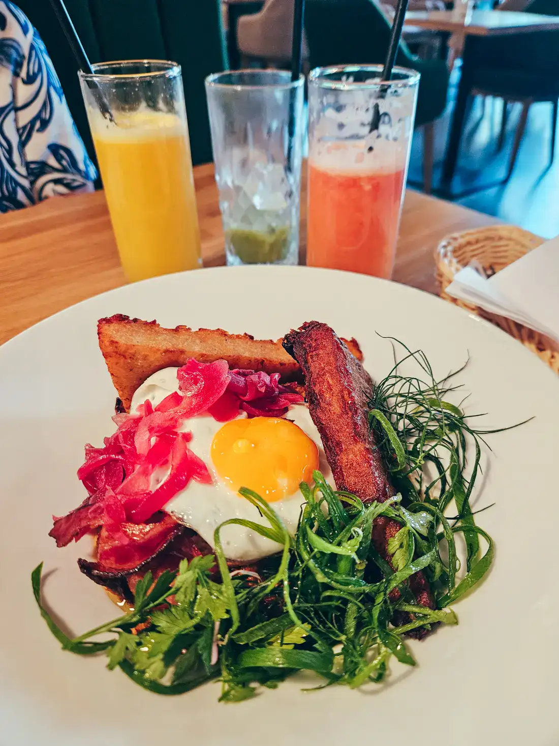 Fried egg with two hash brown, herb salad and oiled red onion on a white, round plate with two juices and an almost empty glass of matcha in the background, at Amsel, one of the best brunch restaurants in Krakow.