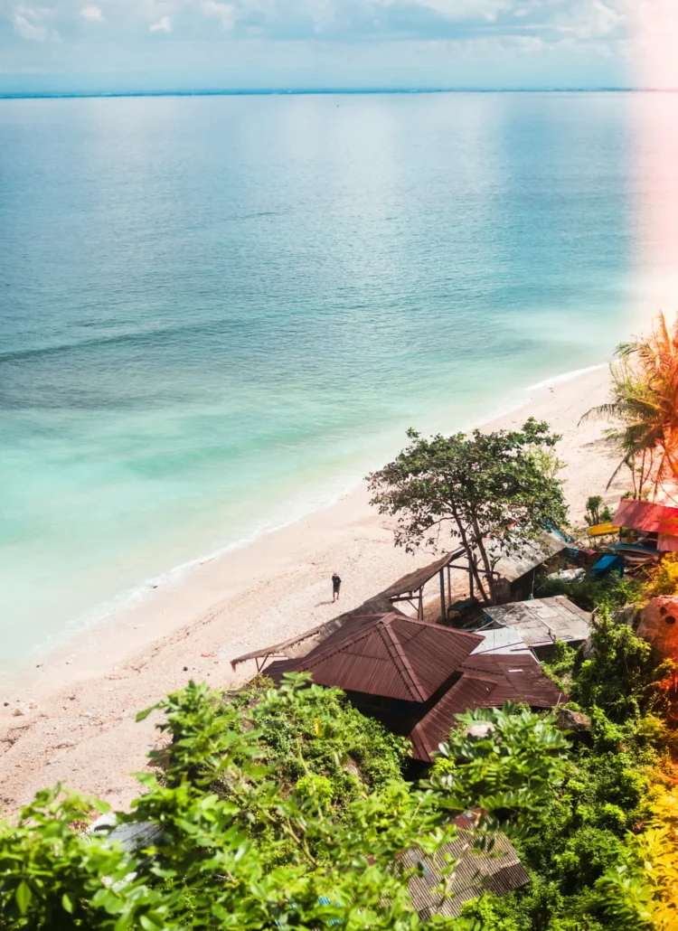 One man walking on the light sand at Thomas Beach lined by palm trees and blue ocean in Bali vs. Lombok.