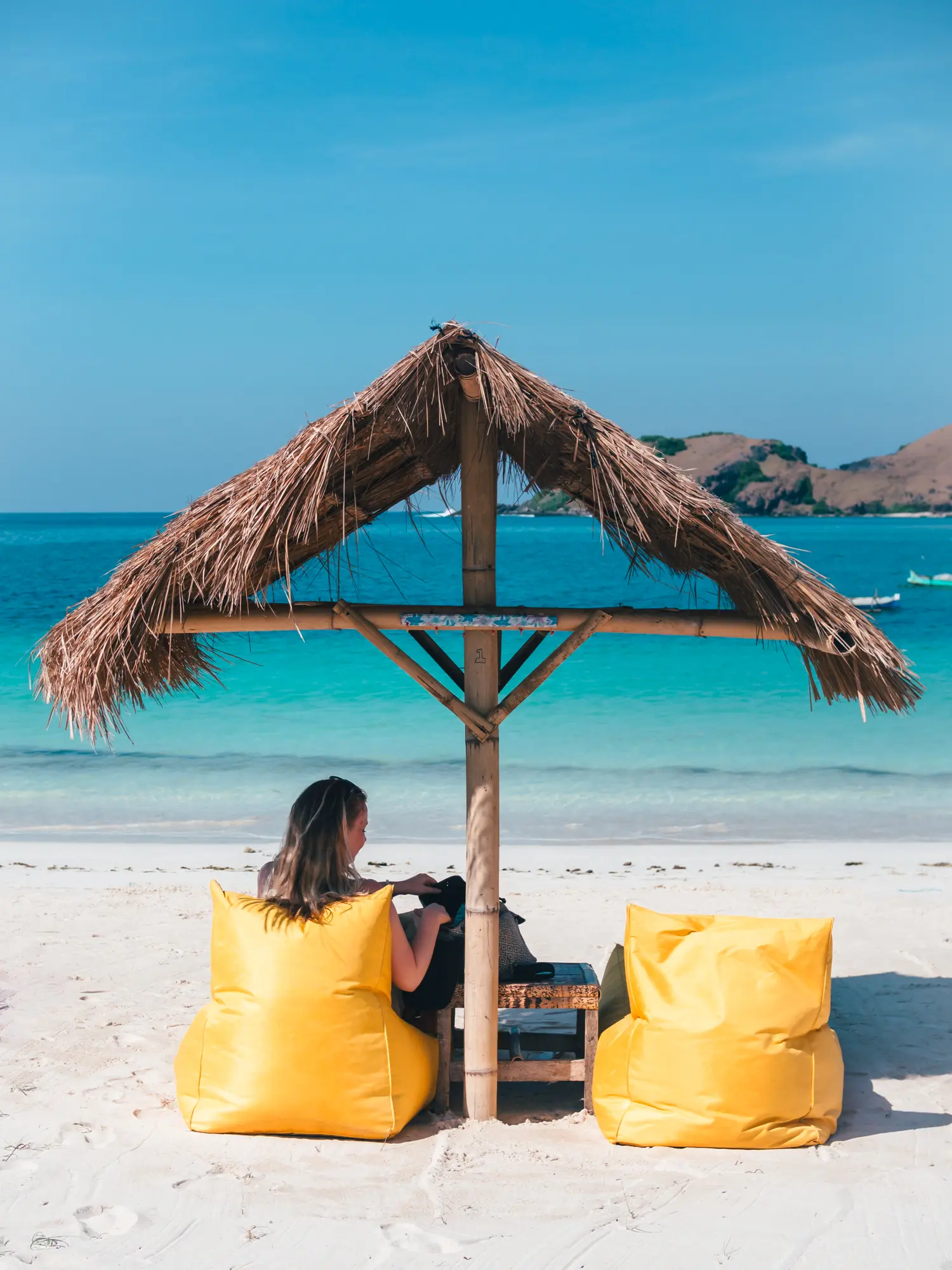 Girl sitting in one of two yellow bean bag under a straw parasol looking out at the turquoise ocean at Tanjung Aan Beach in Lombok vs. Bali.