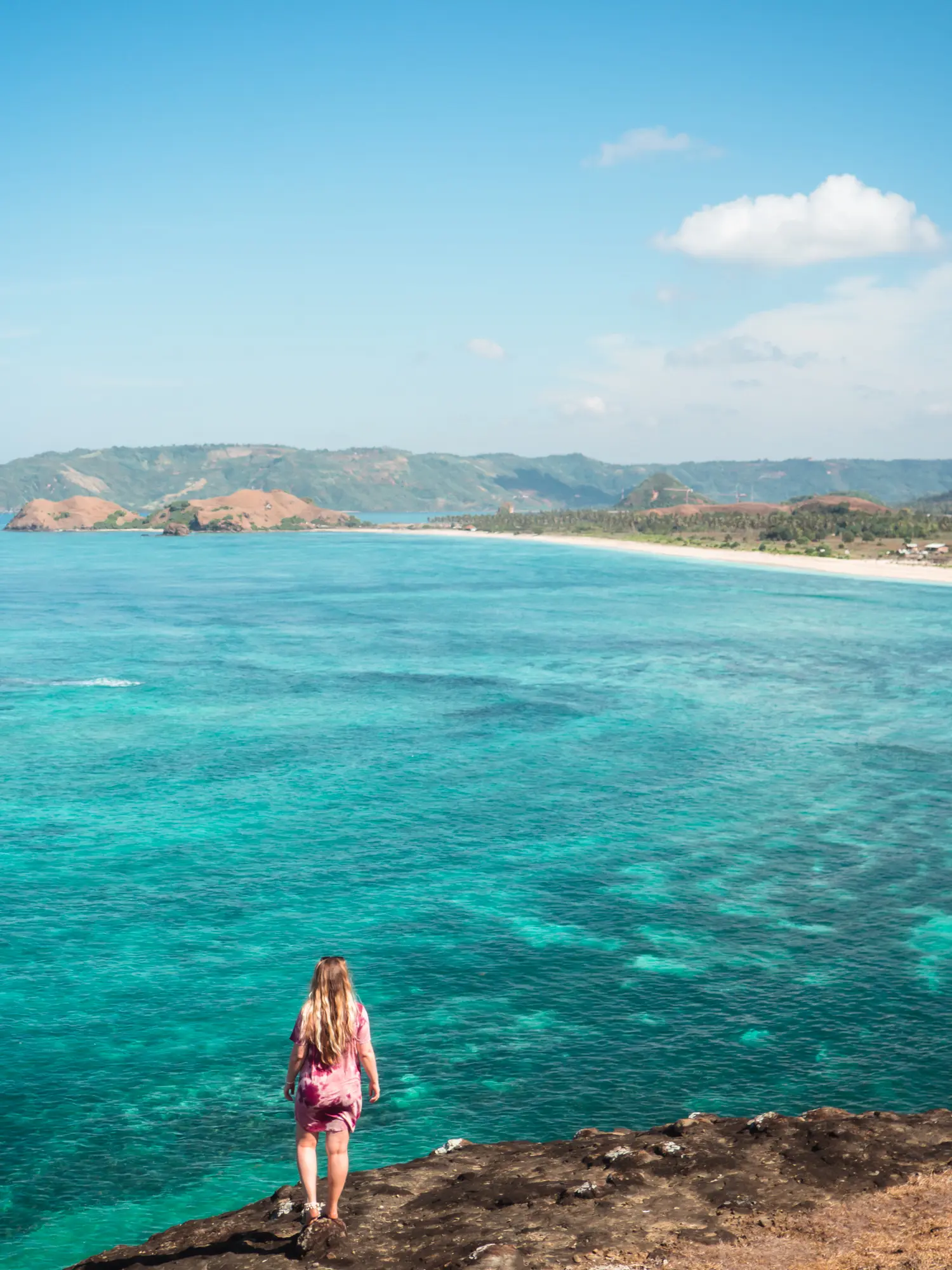 Woman wearing a purple dress standing on the edge of Bukit Merese looking out over turquoise ocean and a beach in the distance. Bali or Lombok?