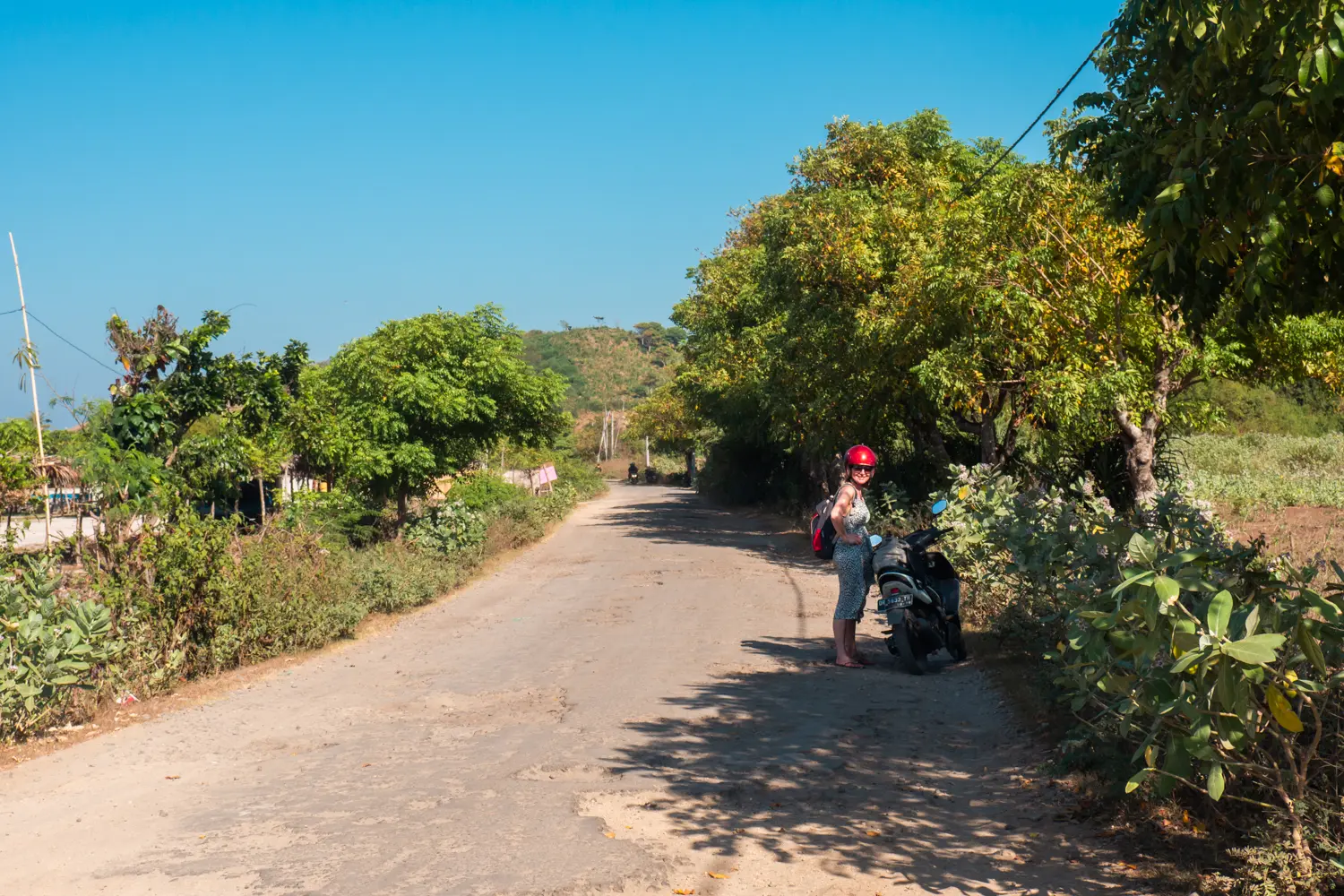 Woman wearing a red helmet standing next to a scooter on the side of a beach road in Lombok vs. Bali.