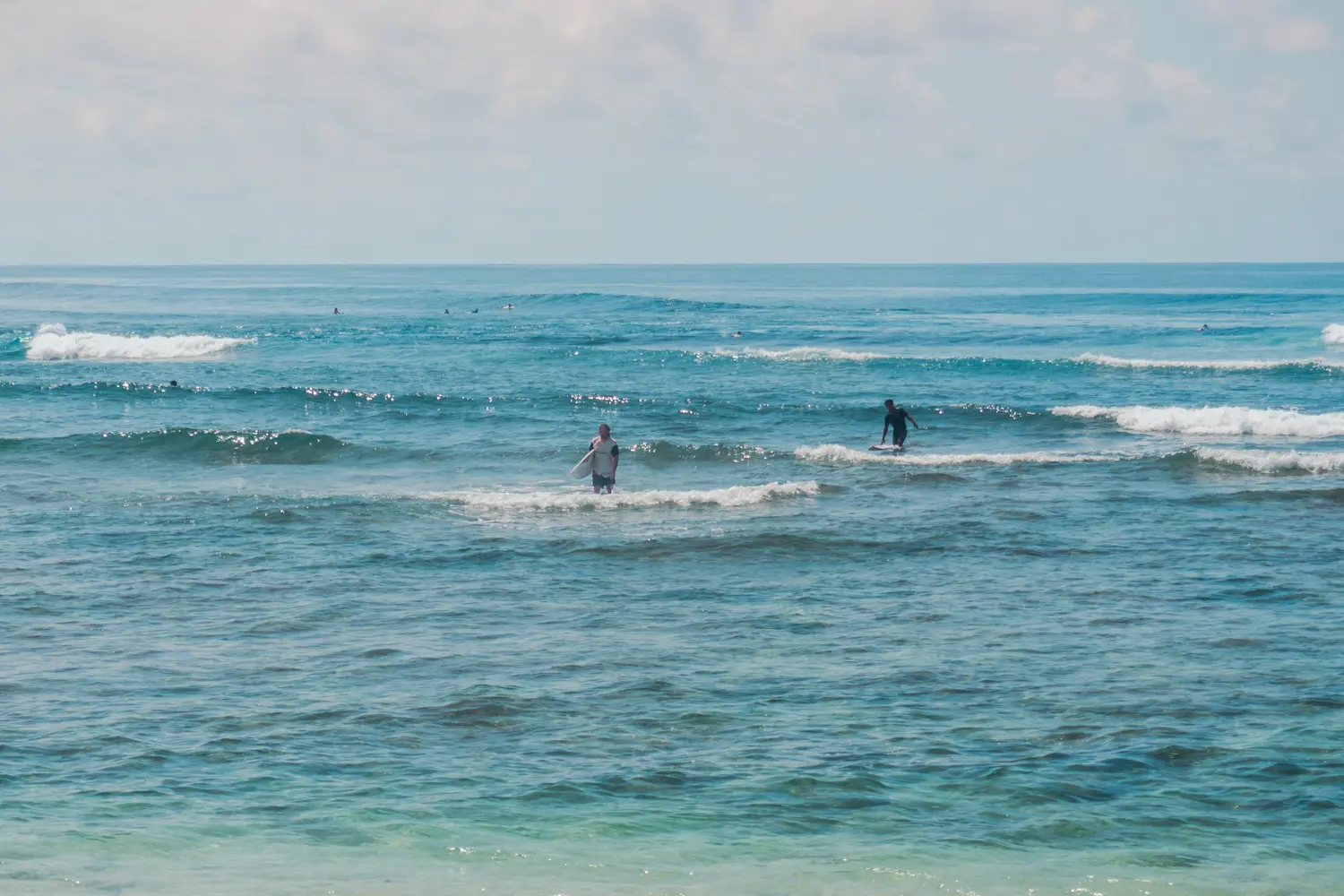 Two surfers coming out of the blue glistening ocean at Green Bowl Beach in Bali. Is Bali or Lombok better for surfing?