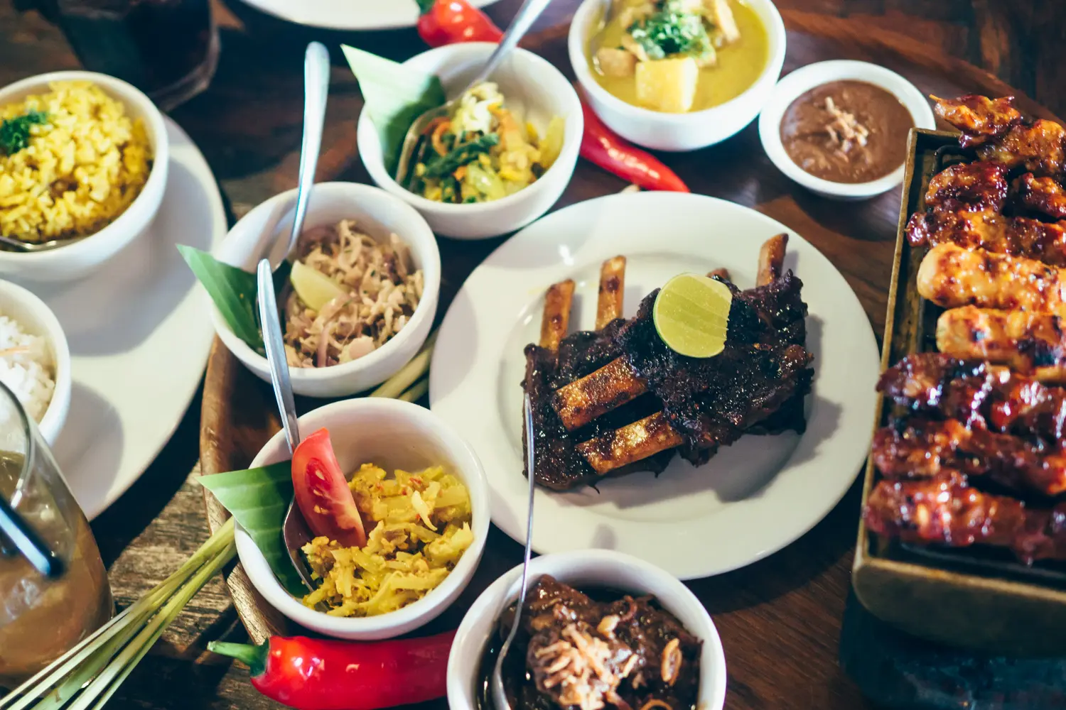 One large white plate of ribs surrounded by large red chilis smaller white plates with Balinese food in Seminyak, Bali vs. Lombok.