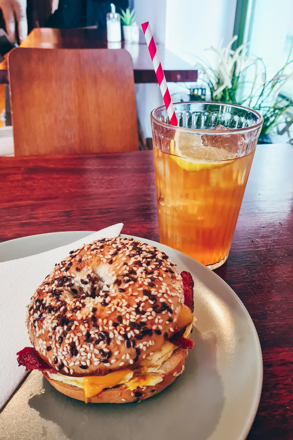 An everything bagel with eggs and bacon on a white plate with an iced tea with a red and white straw in the background at Bagelmama, one of the best places to have breakfast in Poland.