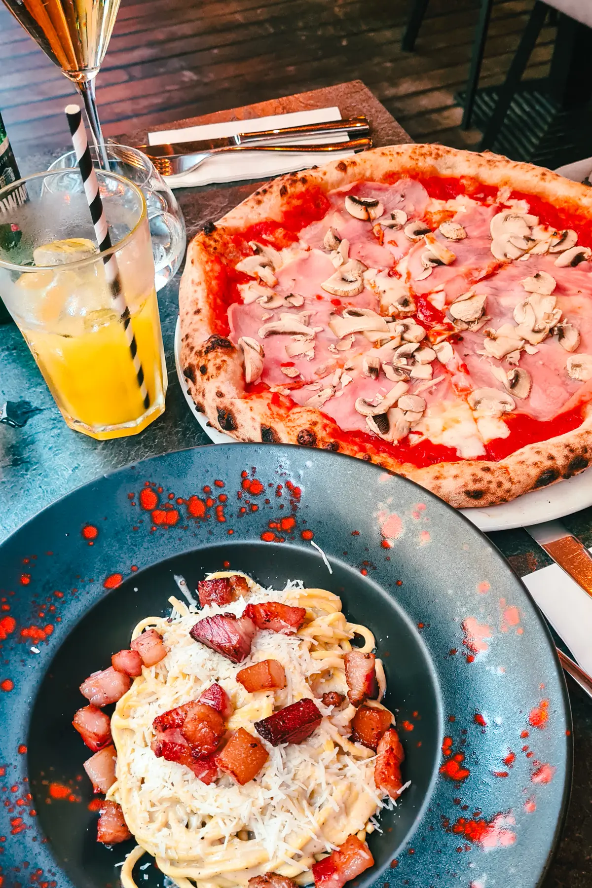 Spaghetti Carbonara in a blue bowl with large chunks of bacon, ham and mushroom pizza in the background with a glass of yellow lemonade at Dolabella Due, one of the best Italian restaurants in Krakow.