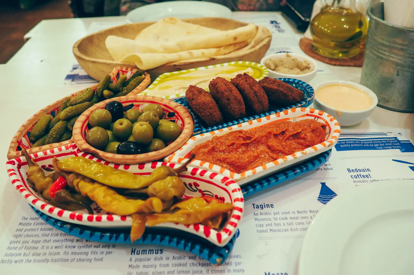 White Hamsa hand plate with red and blue decorations holding falafel, hummus, olives, gherkins and paprikas with bread in the background at Hamsa, one of the best restaurants in Krakow. 