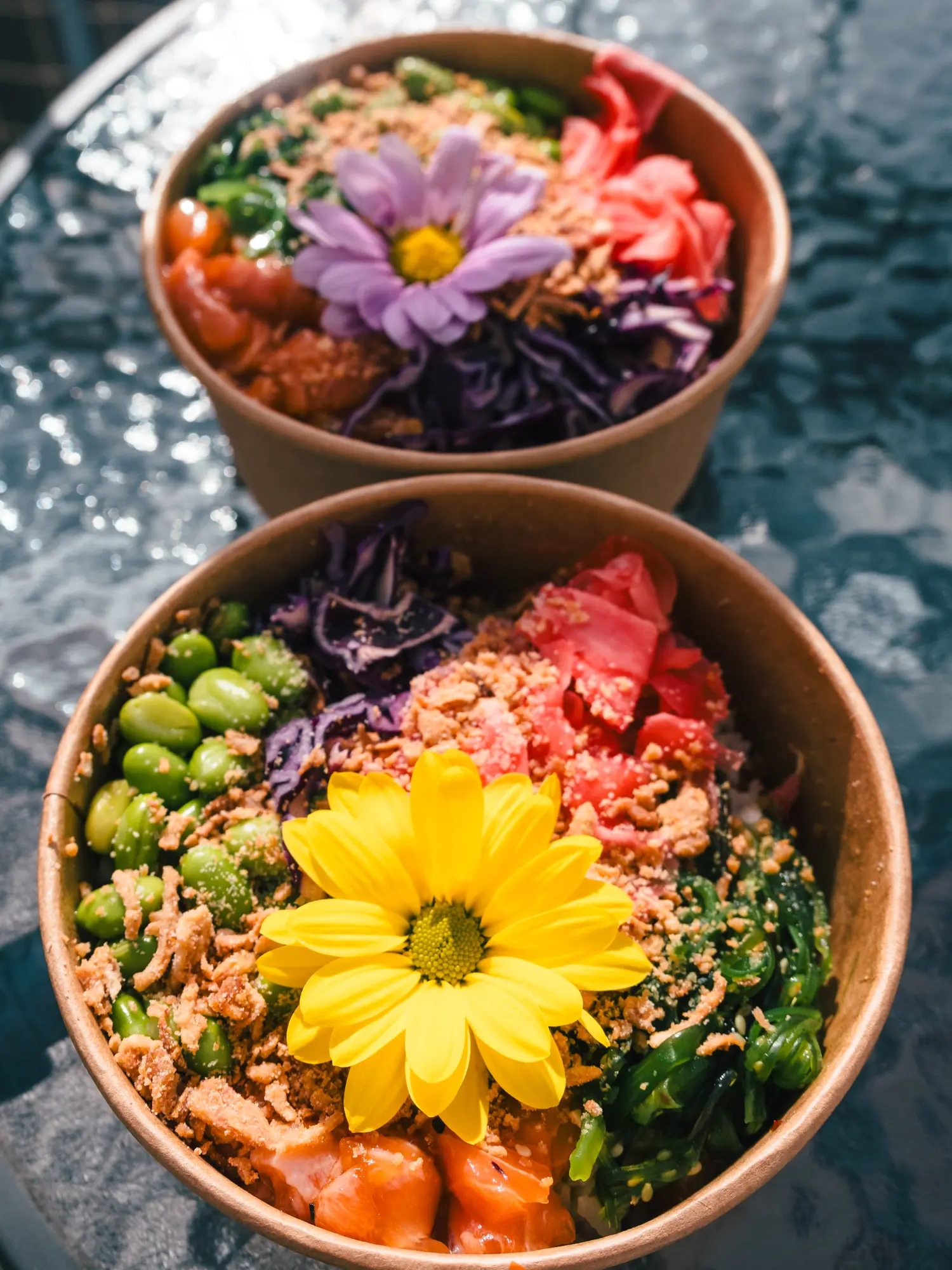 Two poke bowls with edamame, Wakame, salmon, red cabbage, ginger and fried onions, decorated with a yellow and purple flower, from Oké Poké, one of the best restaurants in Krakow.