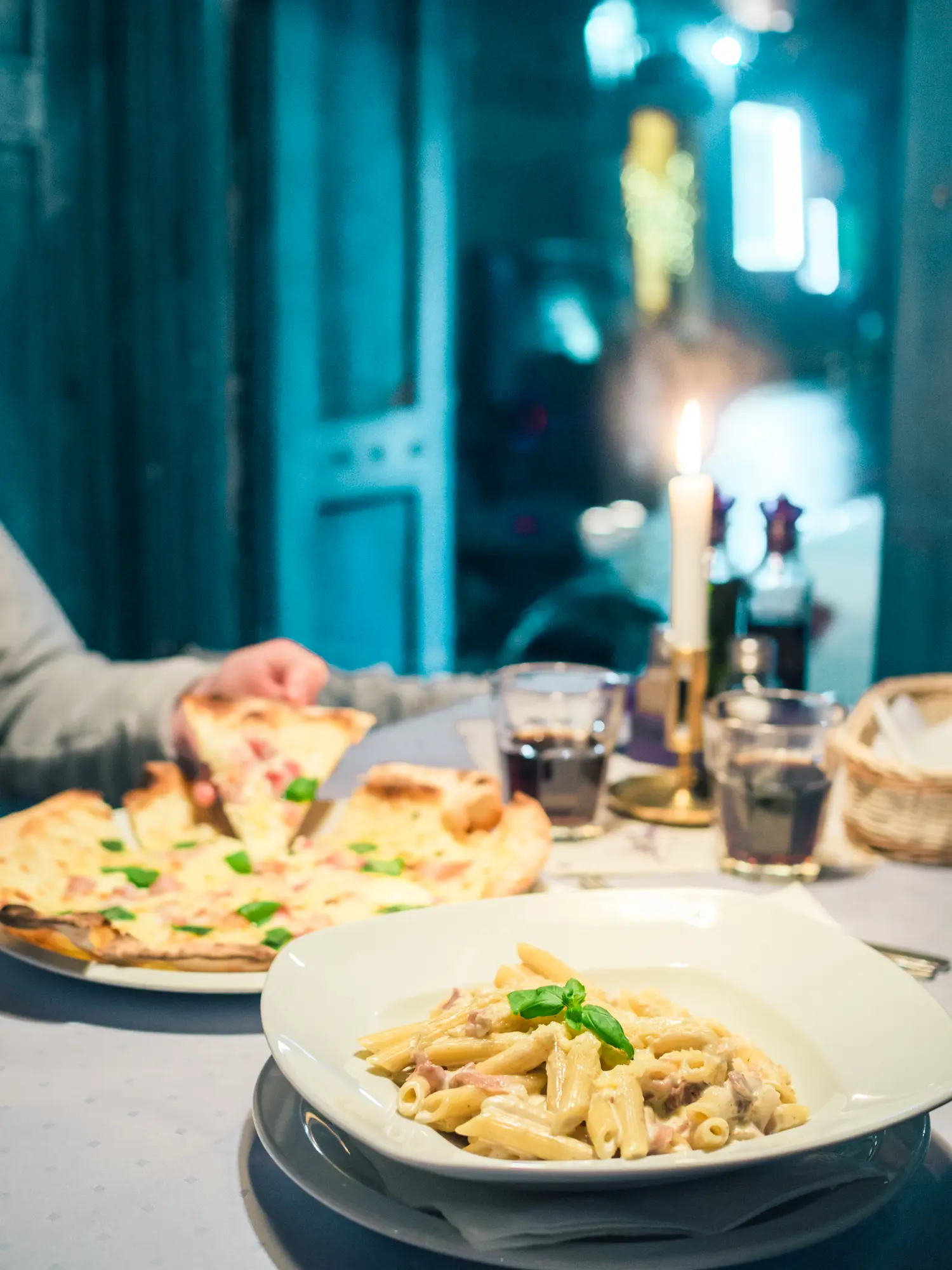 White bowl of penne Carbonara with a pizza and a candle in the background at the blue Trattoria La Famiglia, one of the best restaurants in Krakow.