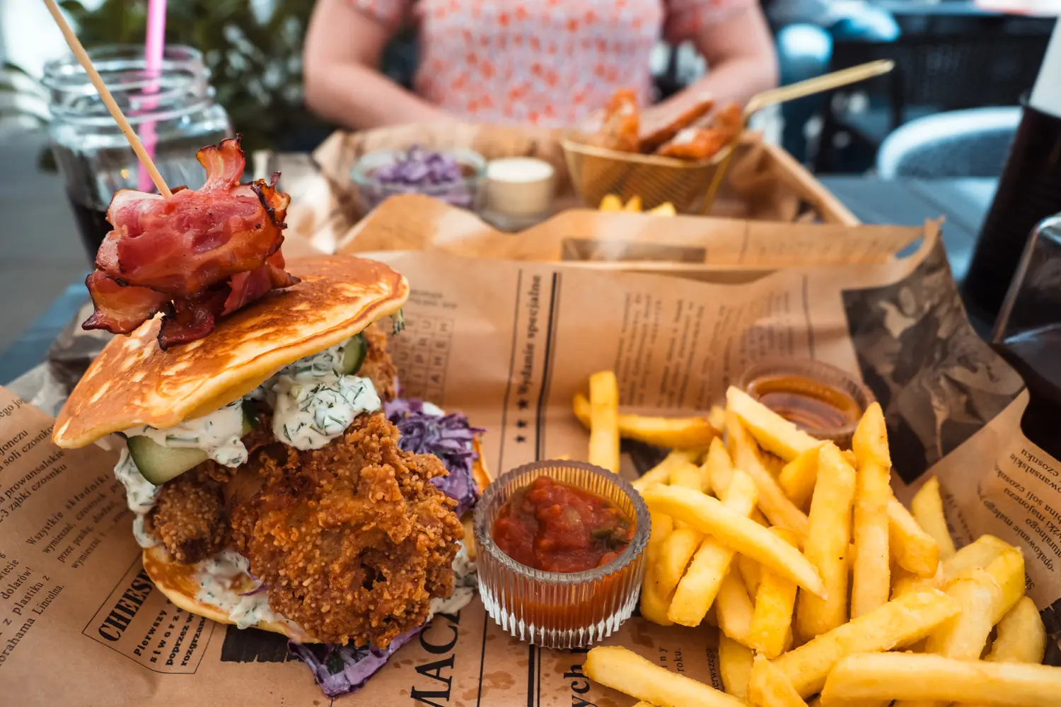 A pancake fried chicken burger with bacon on top and skinny fires with ketchup on a Wooden tray with a beige newspaper at Well Done, the best American restaurant in Krakow.