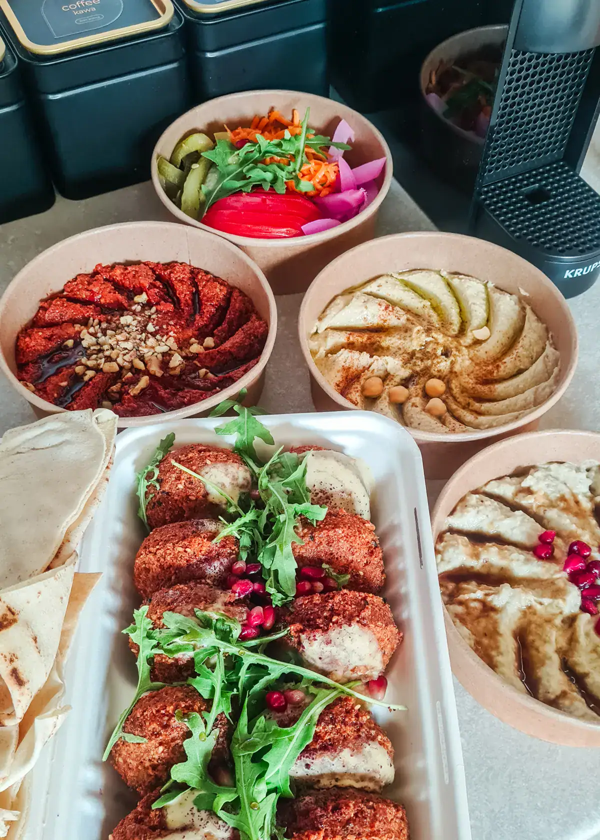 3 round containers of dips topped with oil and nuts, 1 round container with pickled vegetables and a rectangle container with falafel topped with arugula, from Mazaya Vegan Restaurant, one of the best in Krakow.