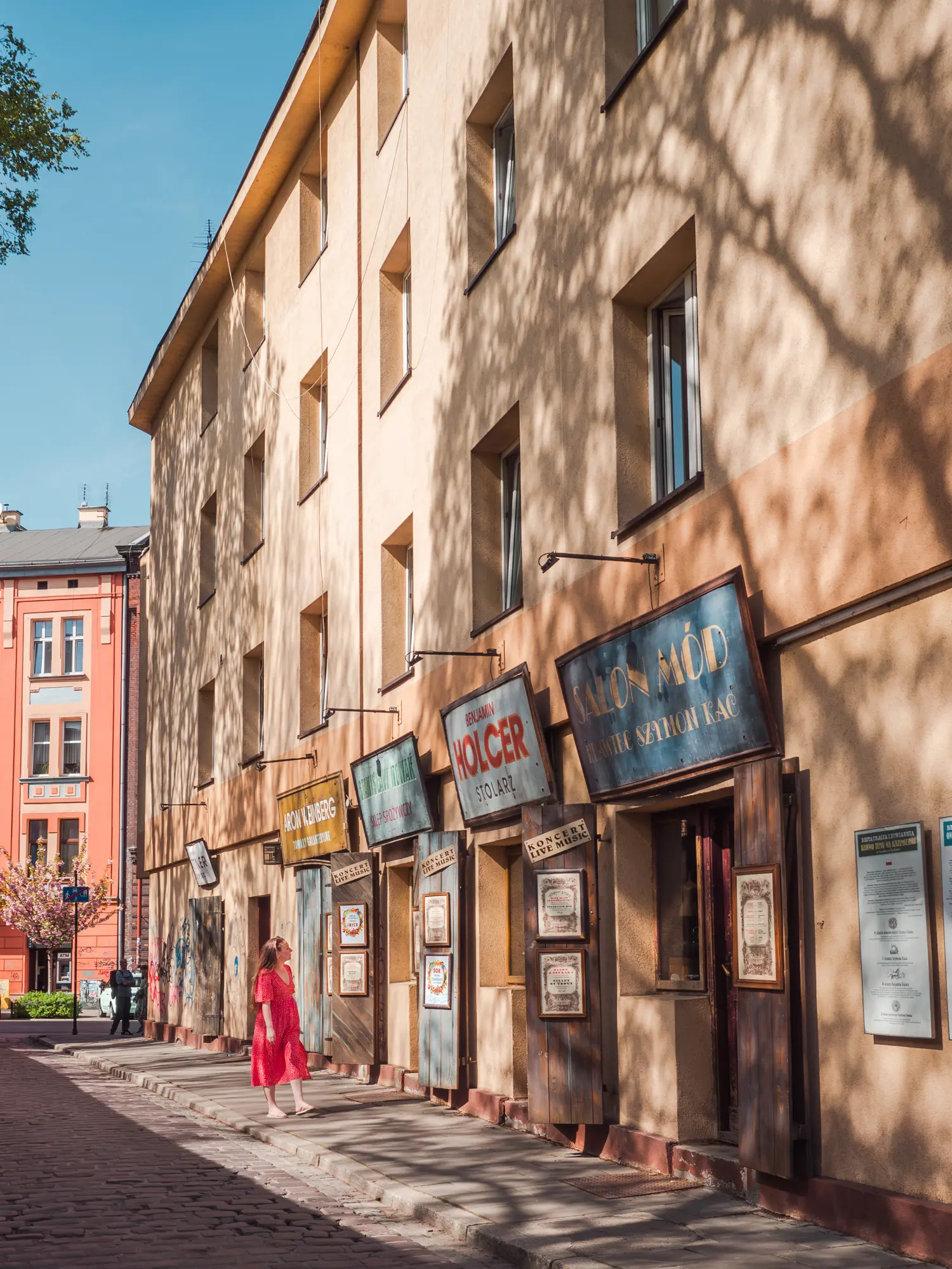 Woman in a red dress walking along a beige building with vintage Jewish restaurant signs in Kazimierz, Krakow.