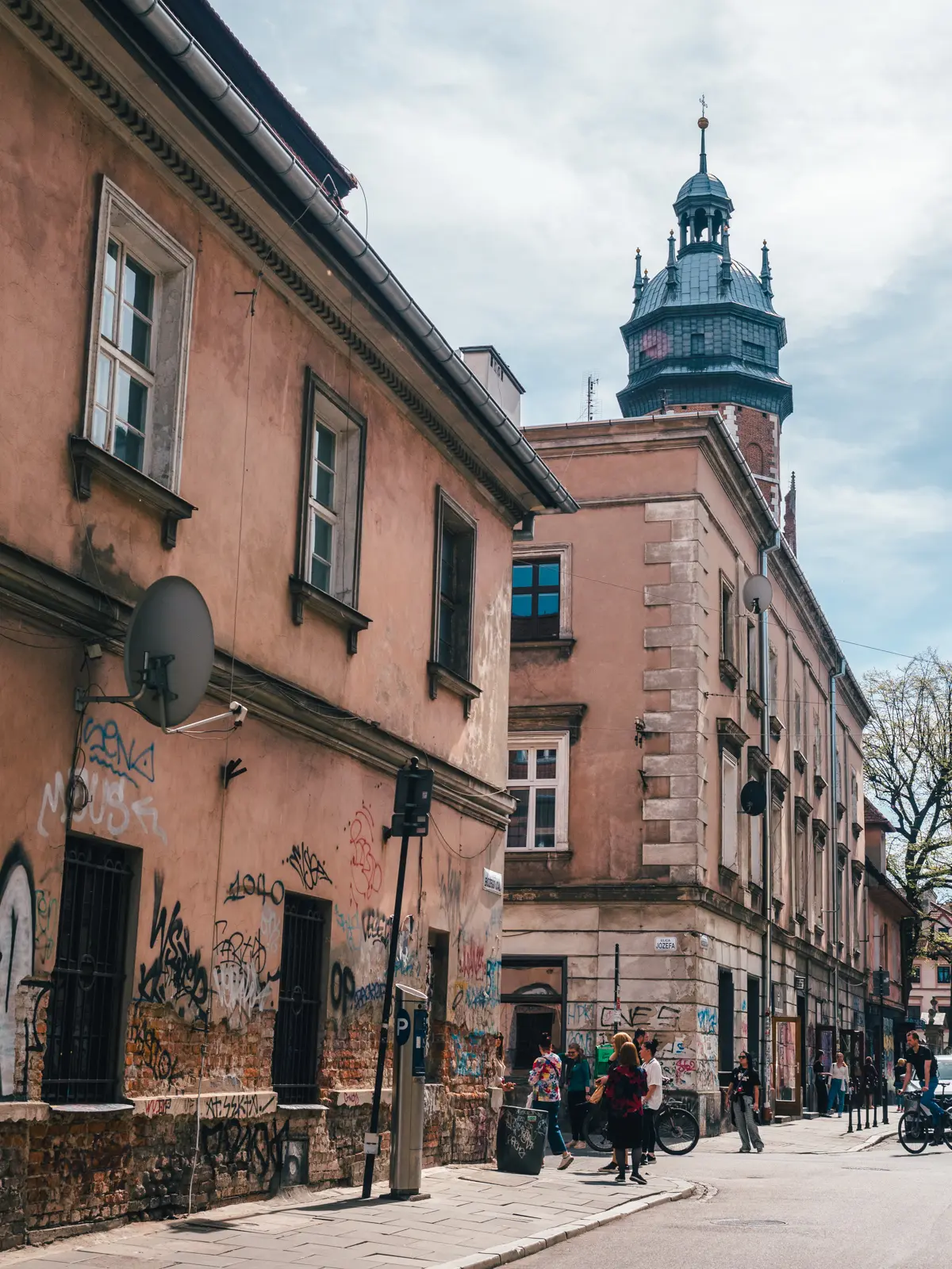 People standing on a typical street corner between two beige buildings with a church tower in the background in Kazimierz.