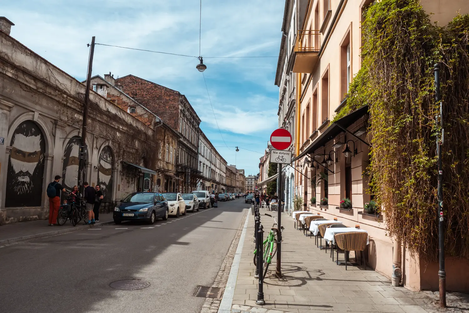 Typical street scene in Kazimierz, one rundown building on the left with graffiti faces, on the right is a restaurants with greenery on the wall and.