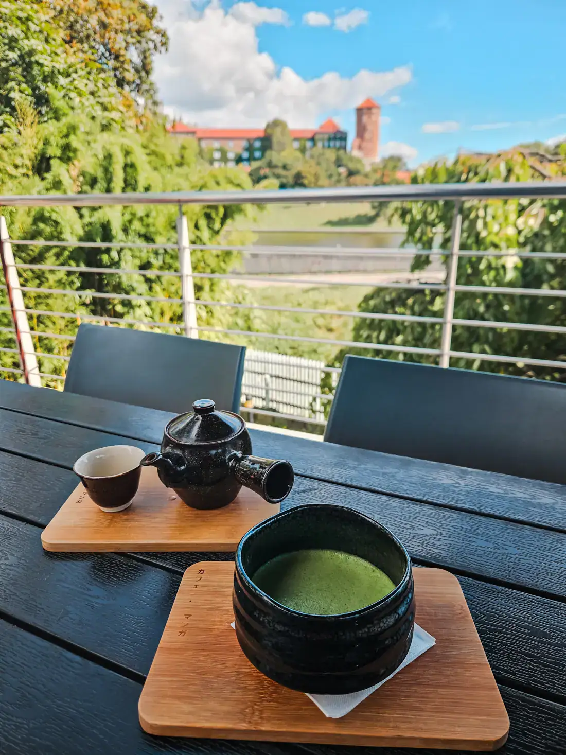 Matcha in a traditional black bowl on a wooden plate, traditional Japanese teapot in the background on a wooden plate, on a black table at Cafe Manggha, with Wawel Castle across the river in the background, one of the best places to eat in Krakow.